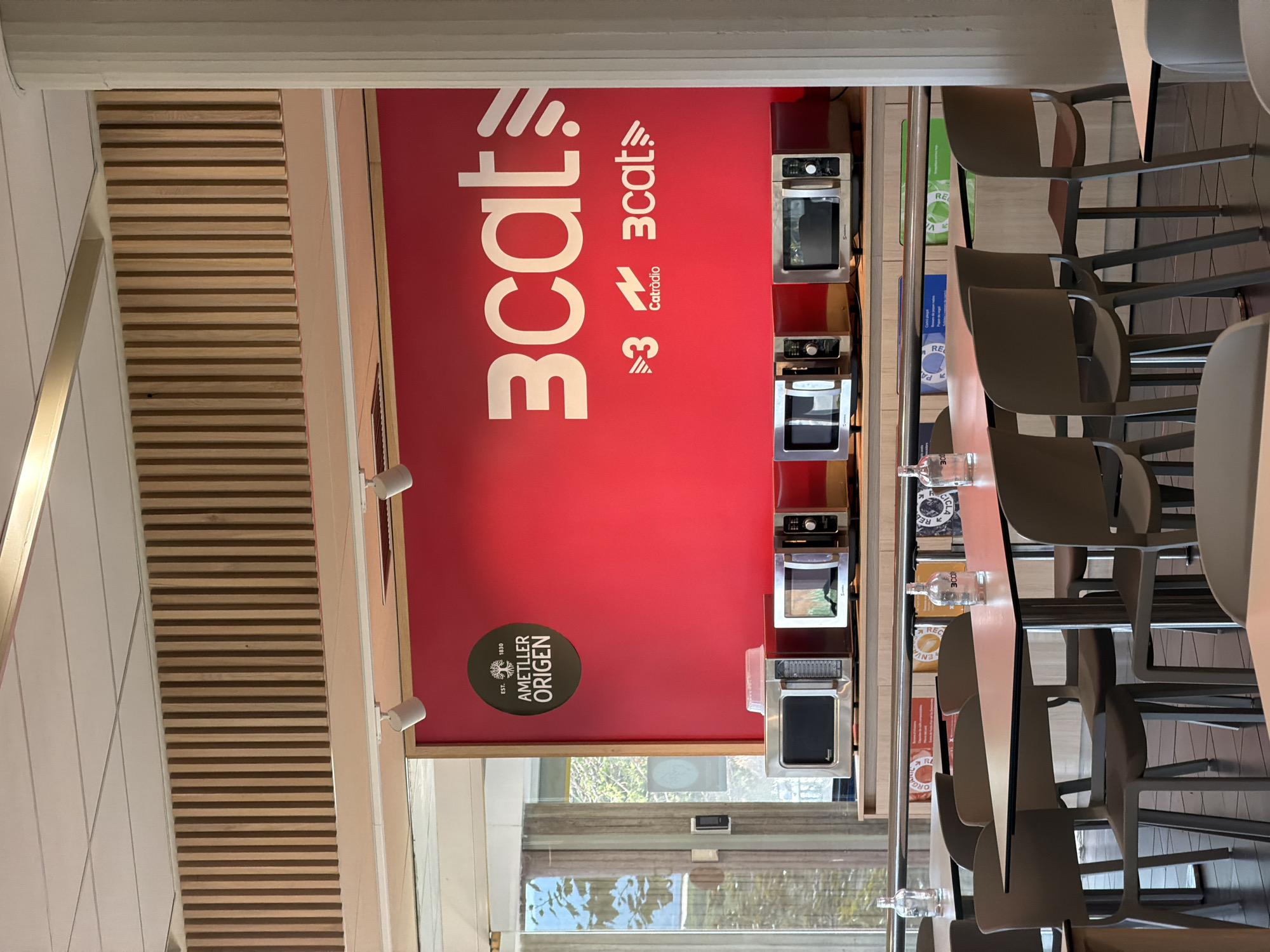 The image captures an interior scene, likely a communal eating area or break room, within the Corporació Catalana de Mitjans Audiovisuals (CCMA) building in Sant Joan Despí, Spain. The prominent feature is a vibrant red wall adorned with the "3CAT" logo in large white lettering, accompanied by smaller logos for "3" and "Catràdio." Below the wall, a row of several silver microwave ovens is displayed on a counter.
To the left of the red wall, a circular emblem with "AMETLLER ORIGEN" and "EST. 1830" is visible. The environment includes tables and chairs, suggesting a functional space for dining or relaxation. In the background, tall, vertical wooden slats form a decorative element on the upper wall. The lighting appears to be artificial, with spotlights visible on the ceiling. The scene is devoid of people, with only tables, chairs, appliances, and wall graphics.