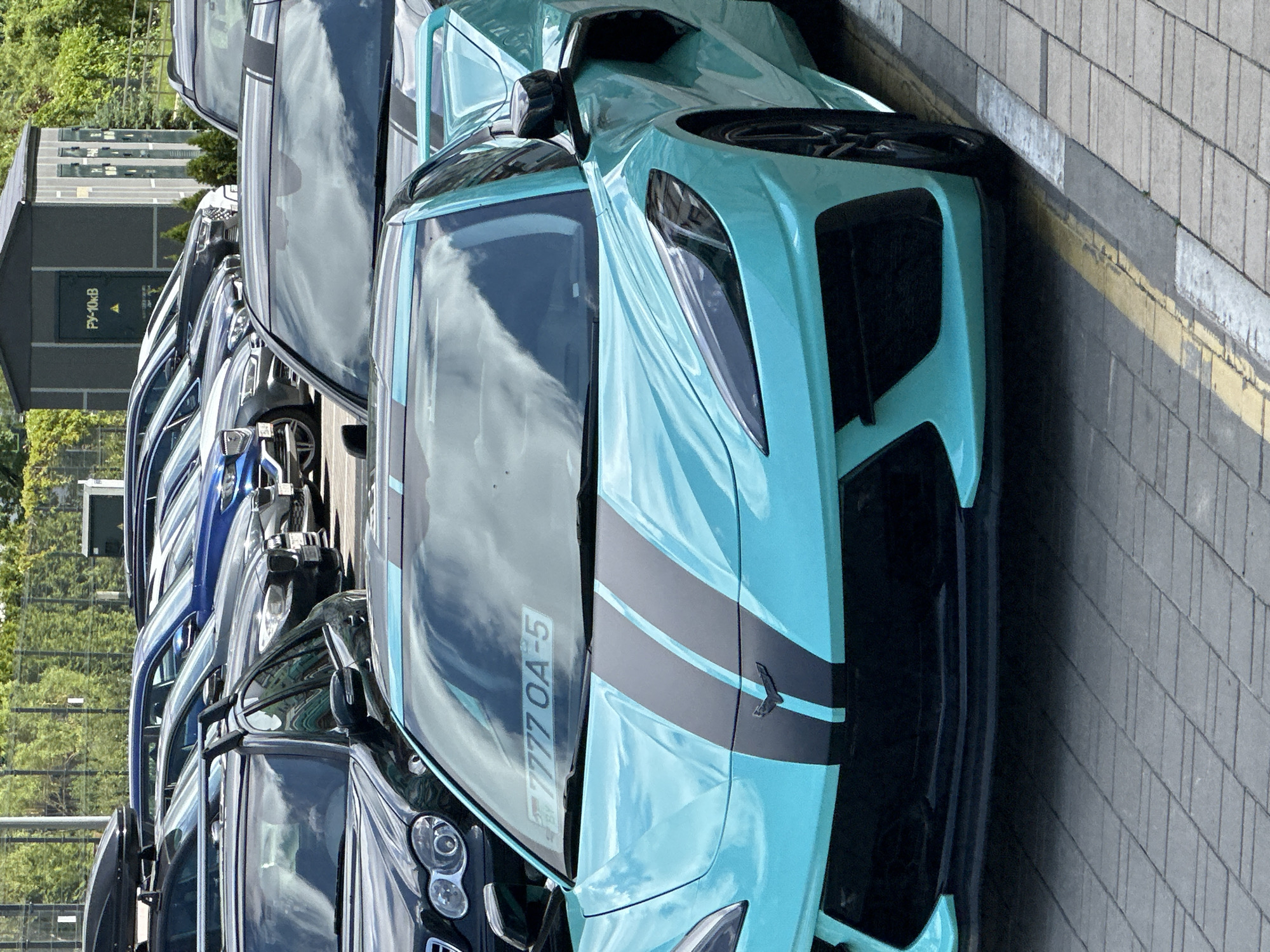 The image depicts a bright turquoise Chevrolet Corvette C8 coupe, featuring dual black racing stripes on its hood, parked in a paved outdoor parking lot. A temporary Belarusian registration plate, "BY 7777 OA-5," is visible inside the front windshield. The car's front fascia includes prominent black air intakes and a lower splitter, with a black Chevrolet Corvette emblem centered on the hood. Black multi-spoke wheels are visible on the front right.
To the immediate right of the Corvette, another vehicle is partially visible, also featuring black racing stripes on its roof. Behind the foreground vehicles, multiple rows of various cars, primarily dark-colored sedans and SUVs, are parked. These include a dark sedan with distinctive circular headlights to the left, and a dark SUV equipped with a roof cargo box in the leftmost background. The parking surface is constructed from light grey paver blocks, with a faded yellow line delineating a parking boundary on the right side of the frame.
In the mid-ground, a green chain-link fence separates the parking area from a dense treeline composed of deciduous and coniferous foliage. Further in the background, a grey, rectangular utility building or electrical substation is visible, displaying the Cyrillic inscription "РУ-10кВ" on its side. The scene is illuminated by natural daylight under a partly cloudy sky, which is reflected in the vehicles' surfaces. The location is identified as a parking area within Бараўлянскі сельскі Савет, Belarus, based on the visible license plate country code and typical infrastructure. No individuals are present in the image.