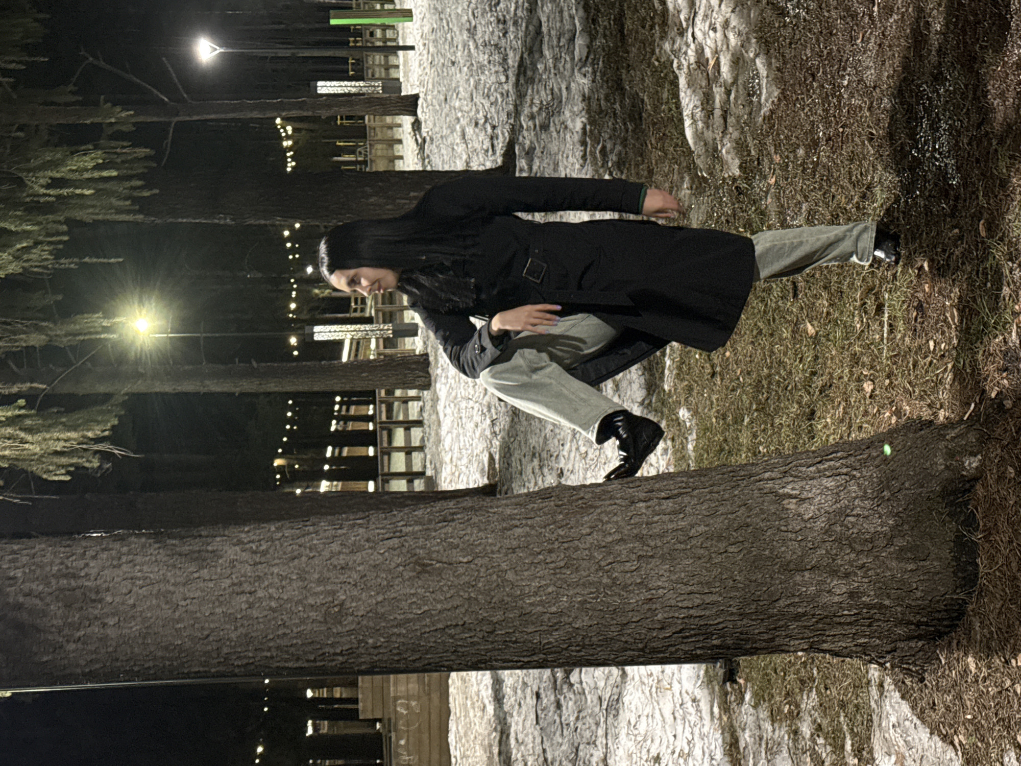 A person is standing on a fallen tree trunk in an outdoor setting, presumably in the city of Votkinsk, Russia. The individual is wearing a dark overcoat, light-colored trousers, and black shoes. The background includes trees, snow, and what appears to be a building illuminated with lights. The scene is captured at night.