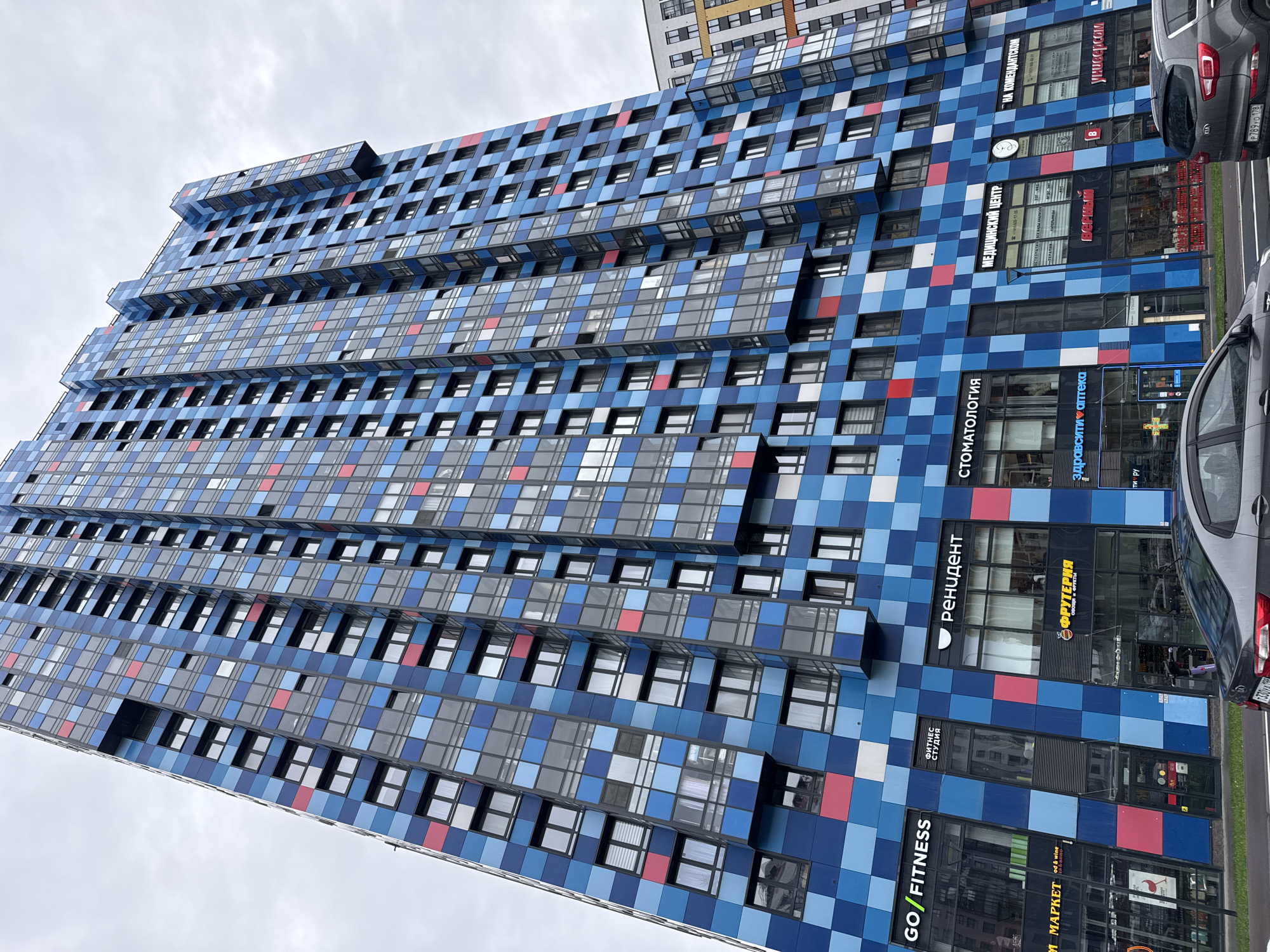 The image shows the exterior of a tall building in Saint Petersburg, Russia. The building's facade is decorated with a grid pattern of blue, white, grey, and red square panels. It appears to be a mixed-use building, with commercial spaces on the lower floors including a fitness center ("GO/FITNESS"), a fruit store ("ФРУТЕРИЯ"), a pharmacy ("здравсити аптека"), a dentist ("СТОМАТОЛОГИЯ"), a medical center ("МЕДИЦИНСКИЙ ЦЕНТР"), and other businesses. Signs are displayed in Russian. The upper floors likely consist of apartments or offices. The sky is overcast. Parts of two cars can be seen at the right, and another building can be seen in the background.
