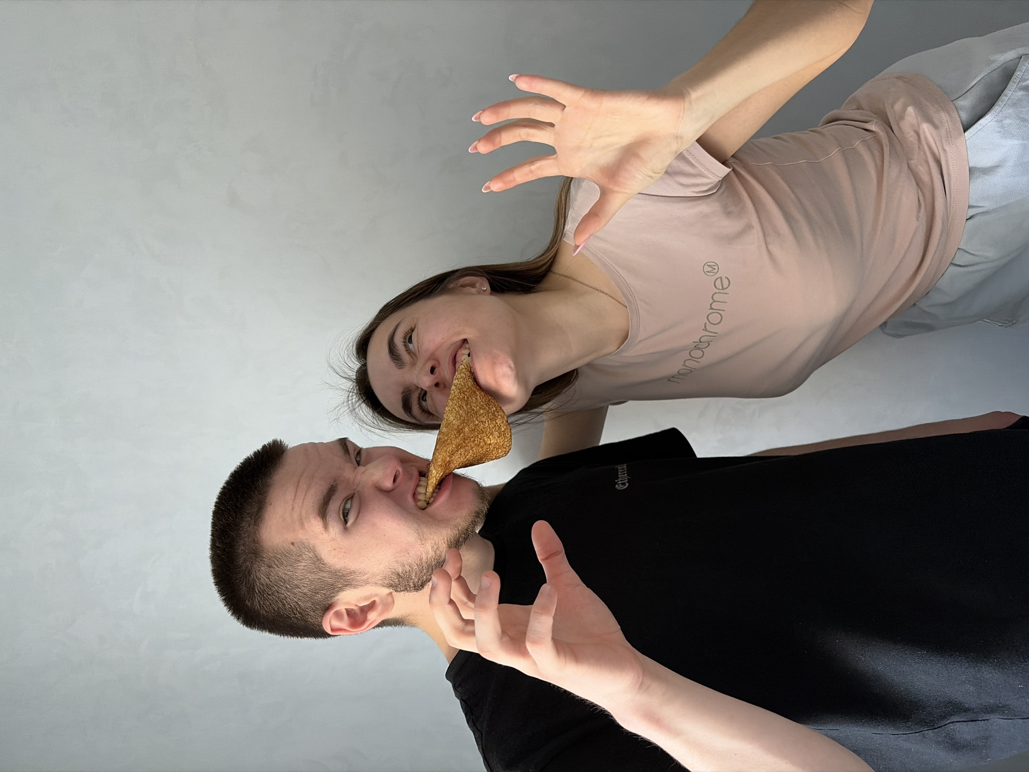 Two individuals are positioned at an undefined indoor location. A male, wearing a black shirt, and a female, wearing a light pink t-shirt, are both attempting to bite into a large, triangular tortilla chip. The male is positioned below the female in the frame. The environment appears to be a room with a plain, light-colored wall.