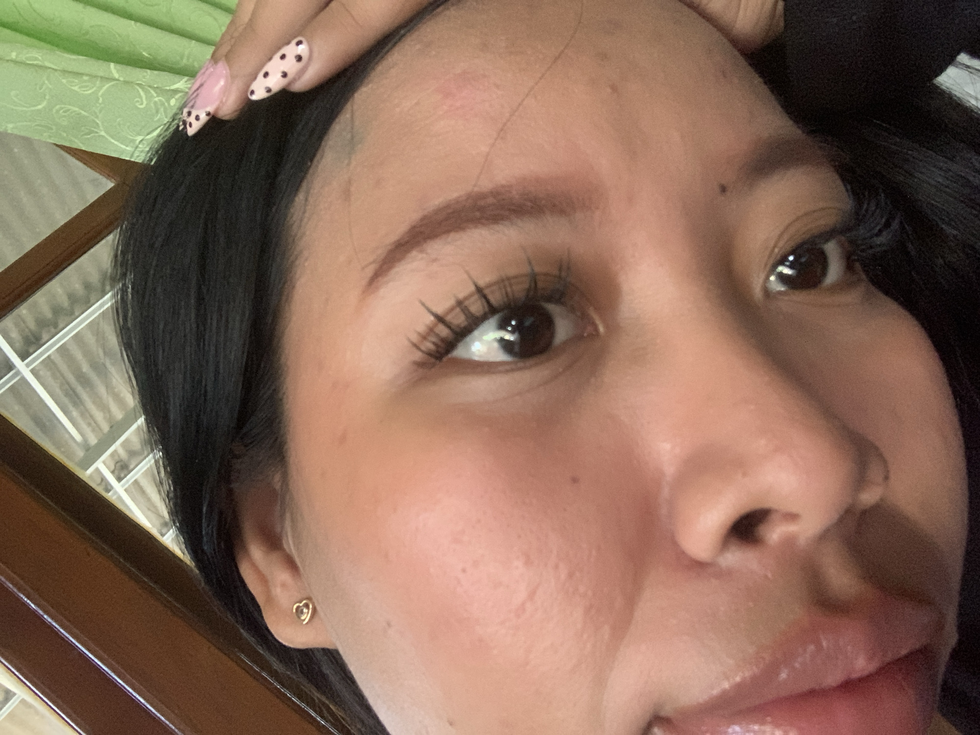 This image is a close-up, high-angle shot of a young woman's face. She has dark hair and dark eyes. She is wearing a heart-shaped stud earring in her left ear and has long, dark eyelashes. Her fingernails are painted pink with dark polka dots and have a heart design. The lighting suggests an indoor setting, possibly near a window with green curtains visible in the background, and a wooden structure. No specific location in Jajag, Indonesia can be determined from the visual information provided.