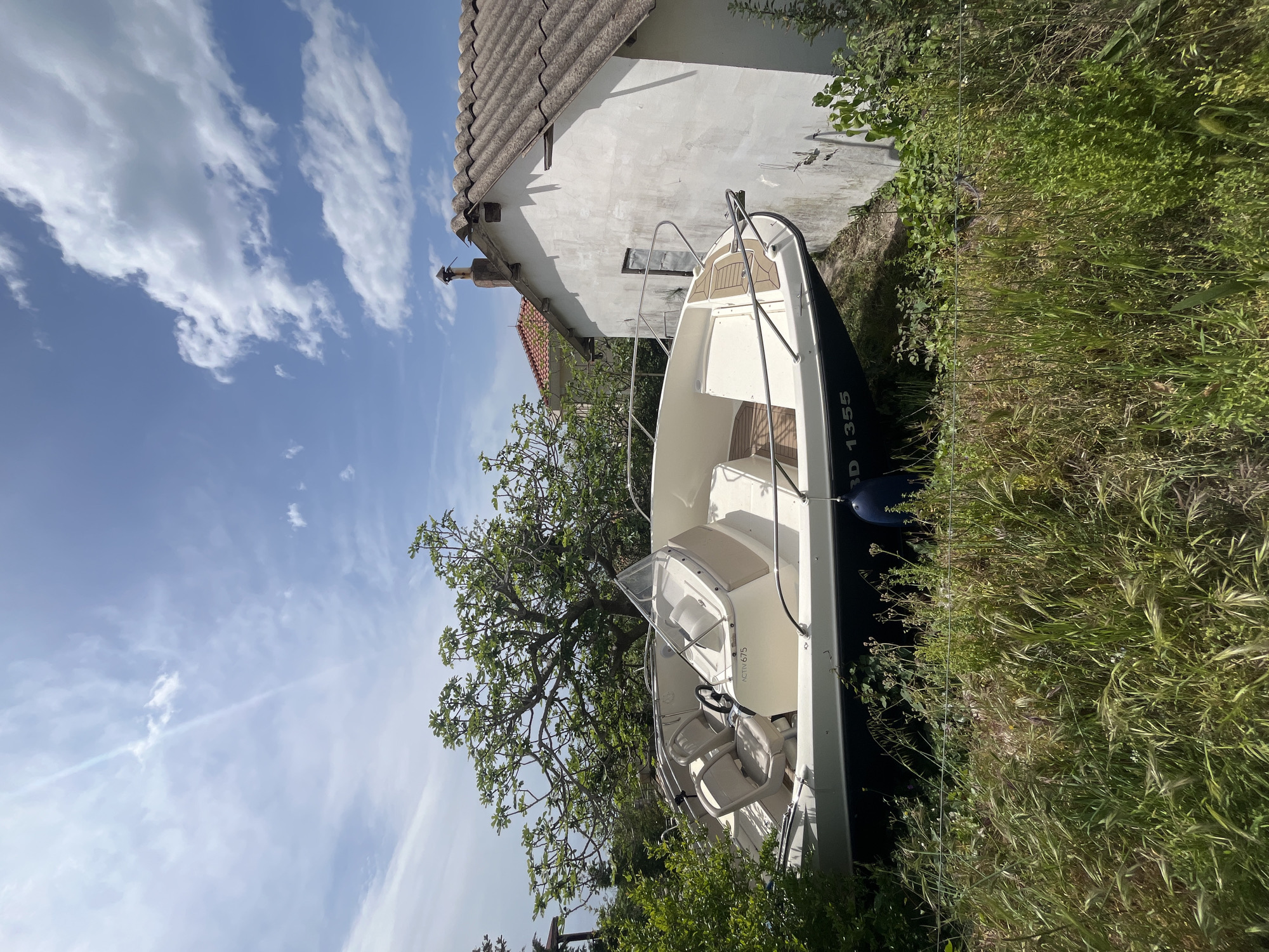 This media file presents a sunlit outdoor scene, featuring a white motorboat stored on land next to a building, surrounded by lush greenery under a clear sky.
The main subject is a white motorboat, likely a recreational vessel, resting on the ground. Its hull is dark at the stern, displaying the registration "RD 1355" in white lettering. The interior of the boat is light-colored, featuring a clear windshield, steering console, and seating with cushions. The text "ACTIVA 675" is visible on an interior side panel. Metal railings are present around the bow.
Adjacent to the boat, on its right, stands a two-story building. It has whitewashed walls and a grey, ridged tiled roof, with a chimney extending upwards. A small, dark-framed window is visible on the side of the building.
The ground around the boat and building is covered in dense, overgrown green grass and weeds. A large tree with a spreading canopy and green leaves, possibly a fig tree, occupies the left side of the frame, partially obscuring the view of the building. The sky above is bright blue with scattered white cumulus clouds, indicating fair weather.
The scene depicts a calm, bright daytime setting, consistent with the location context of Blizikuće, Montenegro. There are no people visible, and no specific activity is taking place, suggesting the boat is in storage rather than in use.