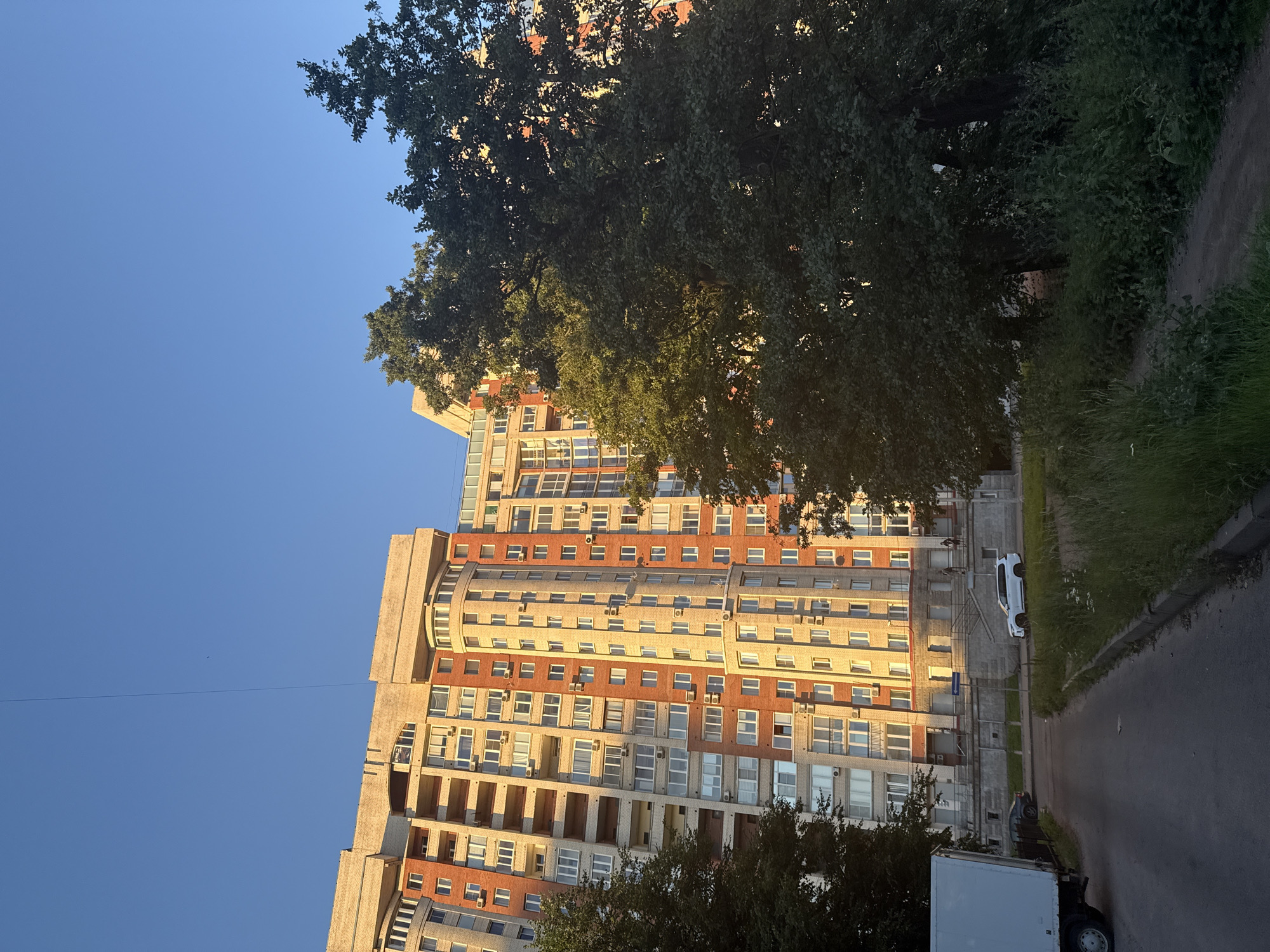 The image depicts a multi-story residential building in Saint Petersburg, Russia, bathed in the golden light of what appears to be either sunrise or sunset. The building, characterized by its light-colored facade with brick accents and numerous balconies and windows, dominates the background. In the foreground, a paved road curves towards the building, with a white truck parked on the left side. A white SUV is visible on the road in front of the building, and several figures can be seen standing near the building's entrance. Lush green trees and vegetation frame the scene, with dense foliage on the right side of the image partially obscuring the building and the sky. The sky is clear and blue.
