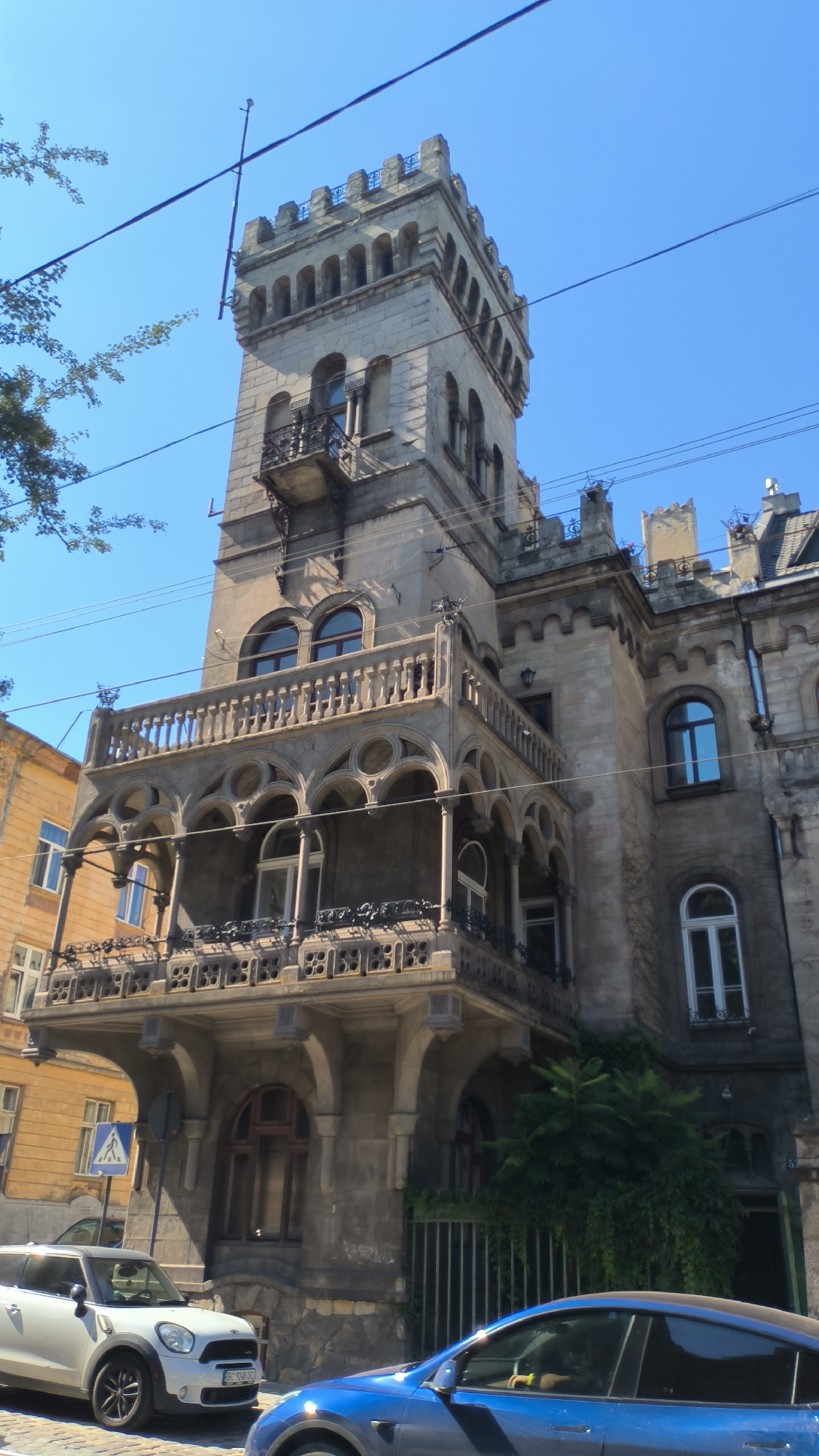 The image captures a side view of an ornate, historic building in Lviv, Ukraine, under a clear blue sky. The building features intricate architectural details, including a prominent tower with battlements, arched windows, and a decorative balcony with a repeating geometric pattern. The facade appears to be made of stone. Adjacent to this main structure, a building with a yellow facade and arched windows is visible on the left. In the foreground, a white mini cooper car and a blue car are parked on a cobblestone street. Electrical wires crisscross the sky above the buildings. Lush green foliage is visible growing against the right side of the main building. The overall scene suggests a daytime setting in an urban environment.