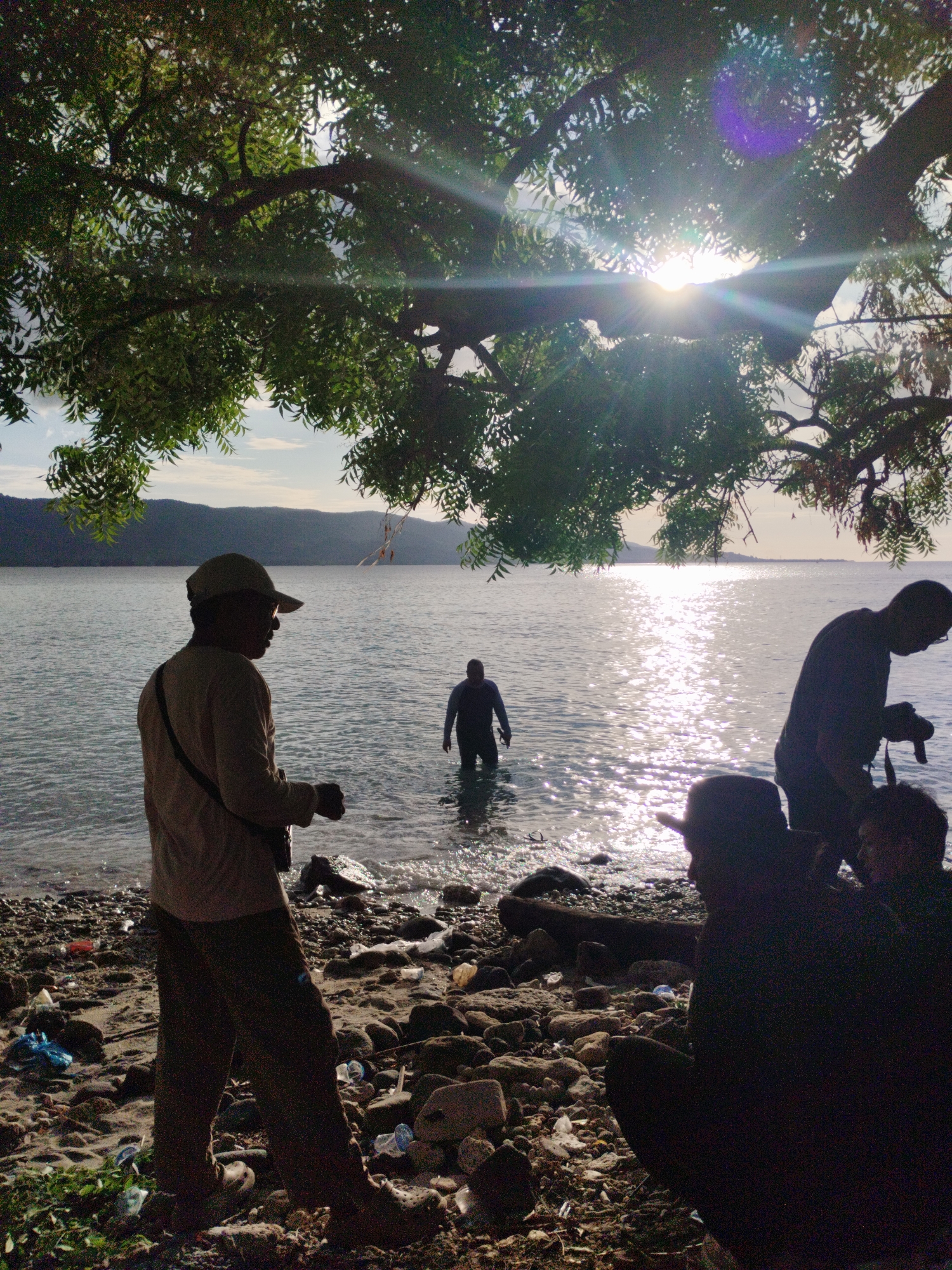 The image shows a coastal scene in Lamreh, Indonesia. In the foreground, a man wearing a cap and casual clothes stands on a rocky beach littered with trash. He is silhouetted against the bright sunlight reflecting off the water. To his right, a person is waist-deep in the water. Further to the right and slightly behind, a man with a camera appears to be photographing. Close to the camera's point of view, two other figures are sitting on the rocky ground, partially obscured by shadows. The background features a bay with hills visible across the water and sunlight is shining through the leaves of a large tree in the upper portion of the frame.