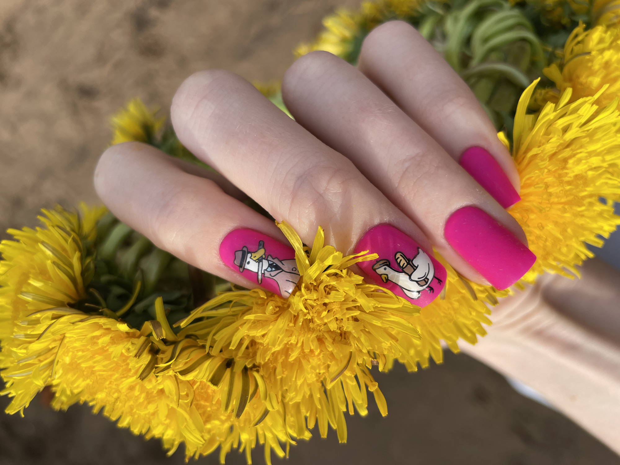 A hand is holding a wreath of dandelions. The nails are painted bright pink. The nails on the index and middle finger have cartoon drawings of ducks, one in a suit and the other holding a baseball bat.
There is no indication of the image being taken in Izhevsk, Russia.