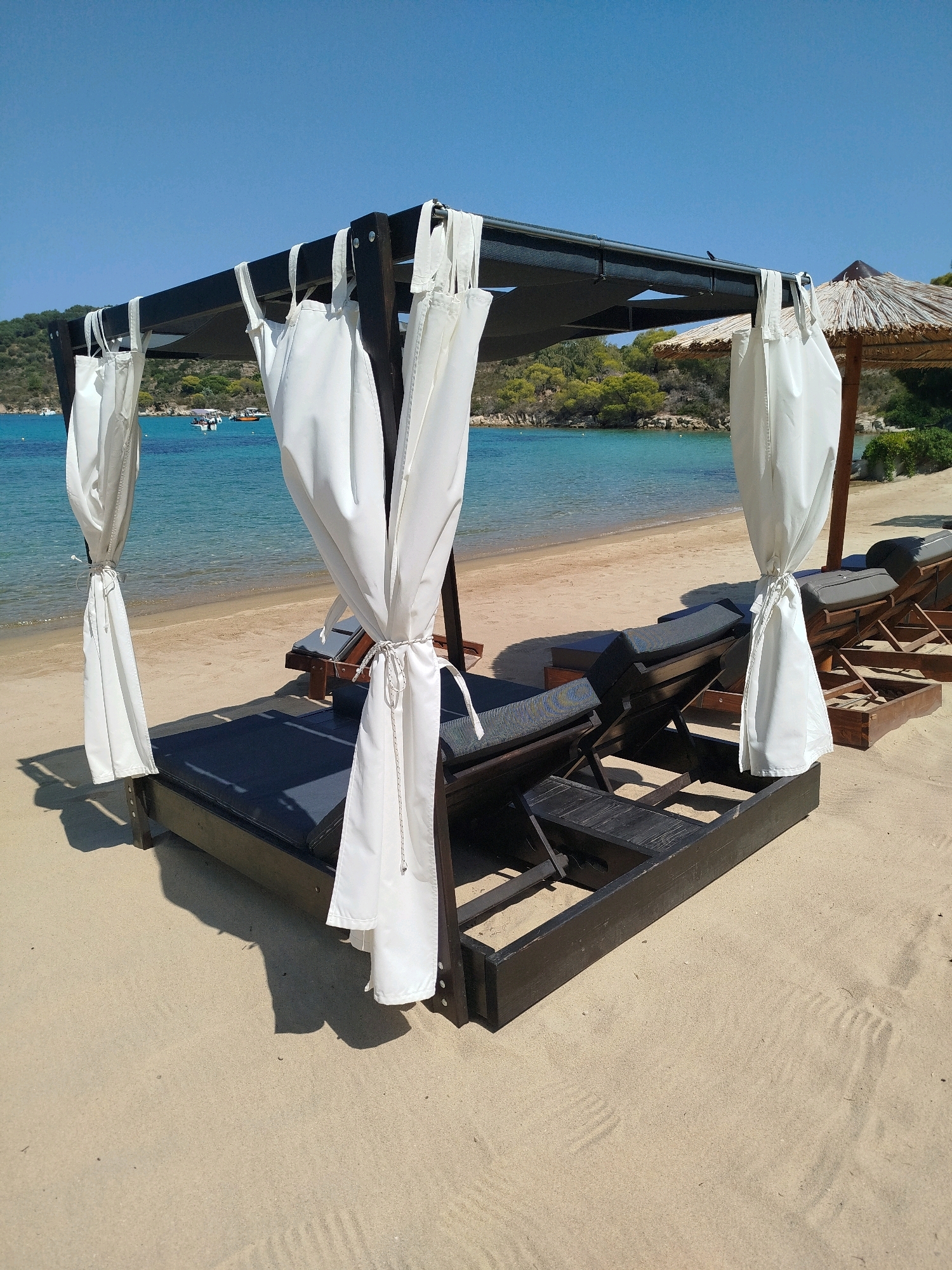 This image depicts a serene beach scene in Sithonia, Greece. In the foreground, a dark wooden cabana-style daybed with white curtains tied back is situated on the sandy shore. The daybed is furnished with a dark grey mattress. Behind it, a row of empty dark wooden sun loungers with grey cushions are arranged, some partially covered by the cabana's shadow. The sea, a clear turquoise color, stretches out to the horizon under a bright blue sky. In the distance, the lush green, hilly coastline of Sithonia is visible, dotted with trees. A few small boats are scattered on the water. The overall impression is one of a peaceful, luxurious beach resort setting.