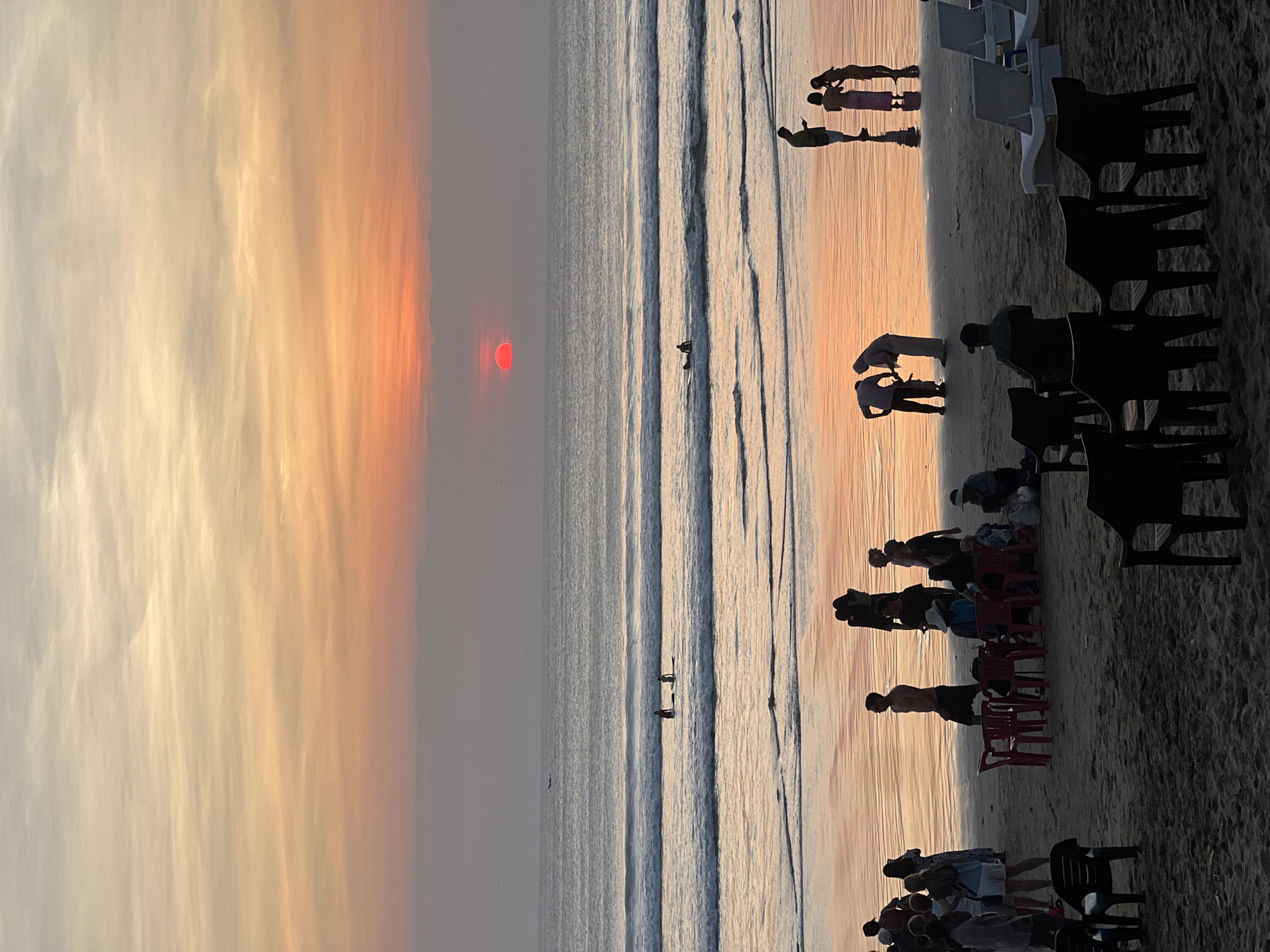 This is a photograph of a sunset on Kuta Beach in Kuta, Indonesia. The sun, a vivid red orb, is partially obscured by a thin layer of clouds and is setting over the ocean. The sky transitions from a soft orange and pink near the horizon to a pale yellow and white at the top. The ocean has gentle waves, and there are several surfers in the water, some standing and some on their boards.
The beach is sandy and wet from the receding tide, reflecting the colors of the sunset. In the foreground, there are numerous people, most of whom are silhouettes against the bright sky. Some are standing in groups, while others are seated on chairs and lounge chairs arranged on the sand. There are also several red plastic chairs scattered around. The overall atmosphere is one of leisure and enjoyment of the natural spectacle.