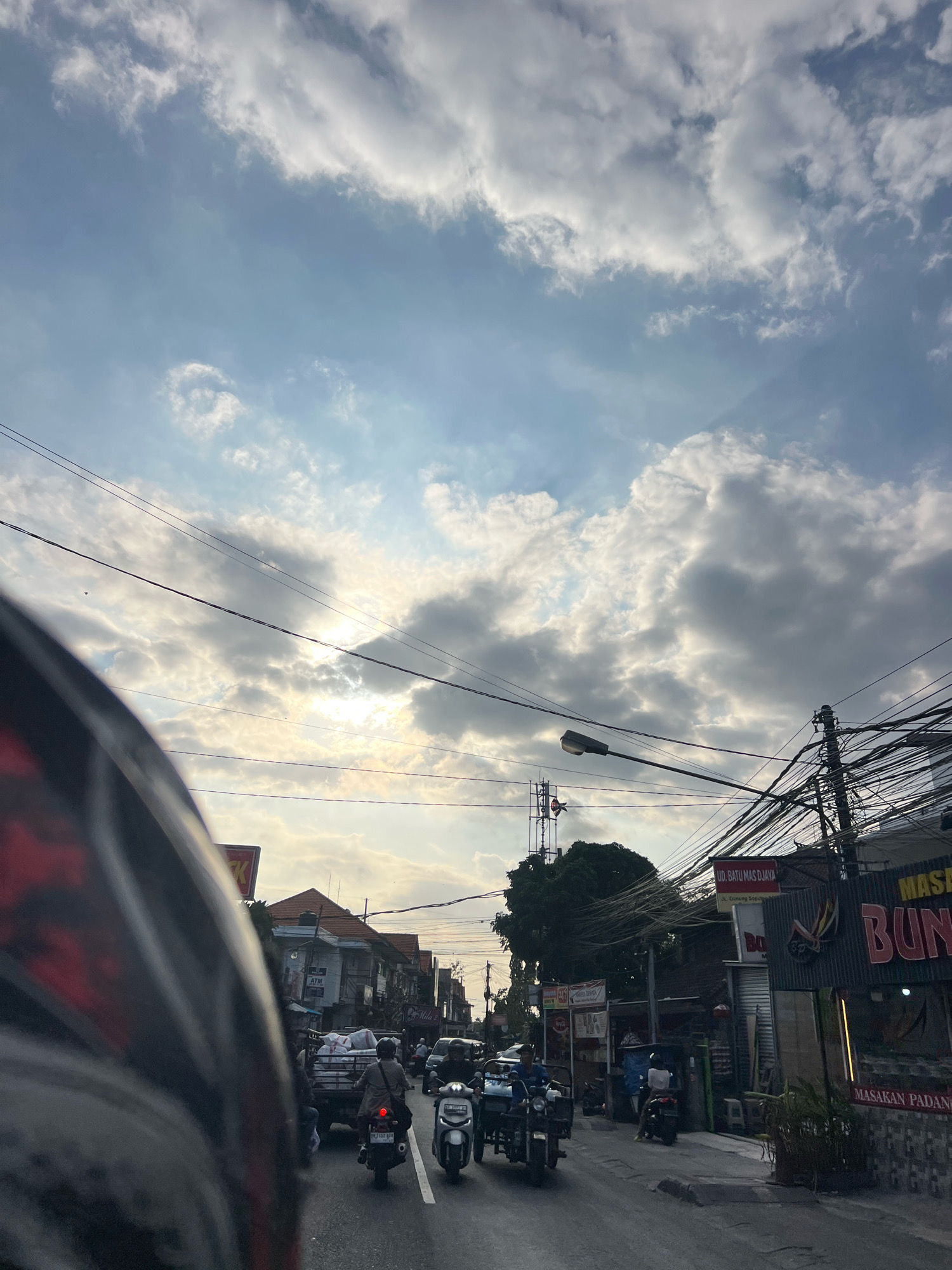 The image captures a street scene in Denpasar, Indonesia, likely during a late afternoon or early evening due to the soft, diffused sunlight. The sky is filled with dramatic clouds, some illuminated by the sun peeking through. Overhead, a dense network of electrical wires is strung across the street, supported by utility poles.
In the foreground, the perspective is from the viewpoint of someone riding a motorcycle, with the side of a motorcycle helmet visible on the left. The street itself is populated by various vehicles, primarily motorcycles, and a few cars and a small truck. People are riding the motorcycles, some wearing helmets, and one person is standing near a building.
On the sides of the street, a row of shops and buildings line the road. Signs are visible, some in Indonesian, indicating businesses such as "MASAKAN PADANG" (Padang cuisine). One building has an ATM sign. The architecture appears typical of a busy urban environment in Indonesia. A cell tower can be seen in the distance behind some trees. The overall impression is of a bustling city street with everyday activity.
