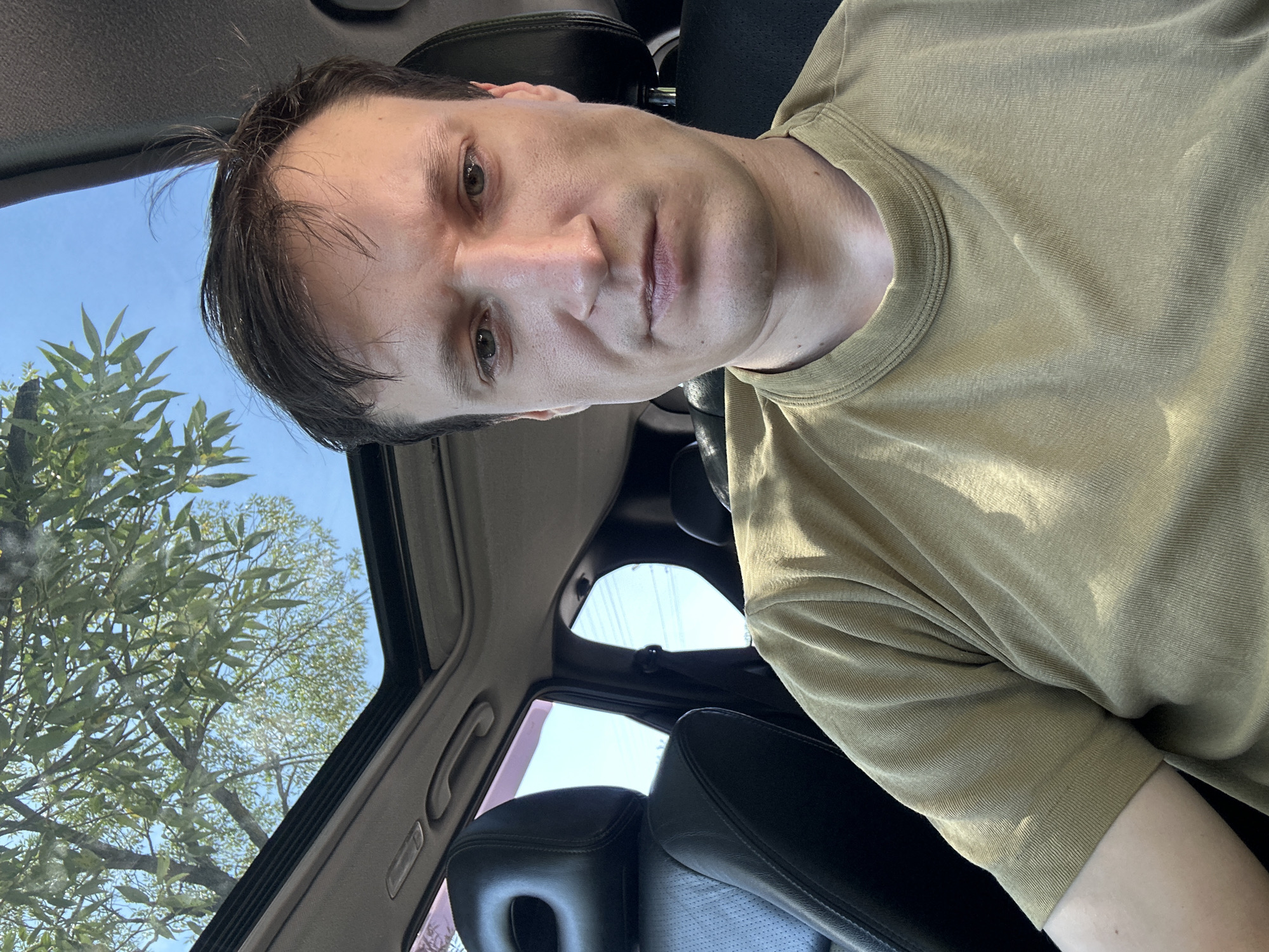 A man is sitting in the driver's seat of a car with the sunroof open, looking at the camera. The car's interior is visible, with black leather seats and headrests. Outside the car, green tree branches and a clear blue sky are visible through the sunroof. The man is wearing a tan t-shirt. The scene appears to be set in an outdoor environment, possibly a street or parking lot, in Makiv, Ukraine, judging by the provided context.