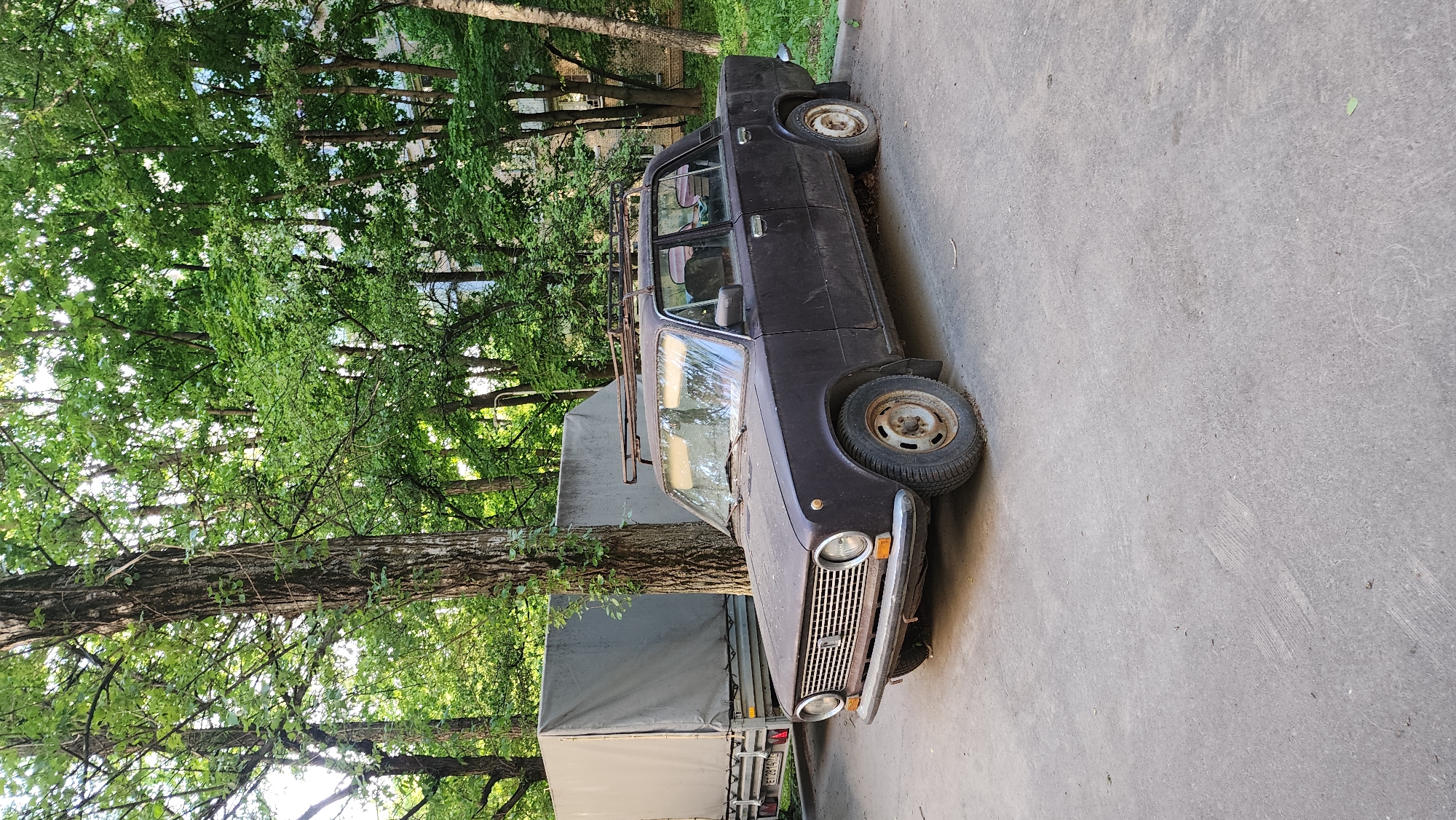 The image shows an old, dark-colored Lada sedan with a roof rack parked on a gray concrete surface. The vehicle appears to be dusty and in disrepair. Behind the car is a light-colored trailer with a covered cargo bed. To the left of the car and trailer are several trees with green leaves. Based on the license plate visible on the trailer (E173XM 777), the location is Moscow, Russia.