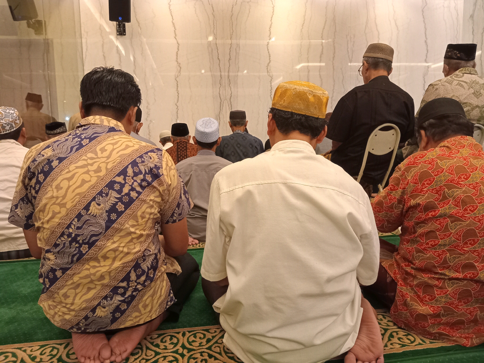 In Randuagung, Indonesia, a group of men are gathered in prayer on a green carpet inside a room. The room's walls are a light color with vertical, darker lines. The men are facing away from the camera, and most are wearing traditional Islamic attire, including various types of caps. In the foreground, two men are in focus. One, on the left, wears a yellow and blue patterned shirt and is kneeling with his feet visible. The other, on the right, is wearing a plain white shirt and is also kneeling. In the background, other men are visible, some sitting on chairs, and others kneeling on the carpet. There's a speaker mounted on the wall above and to the left.