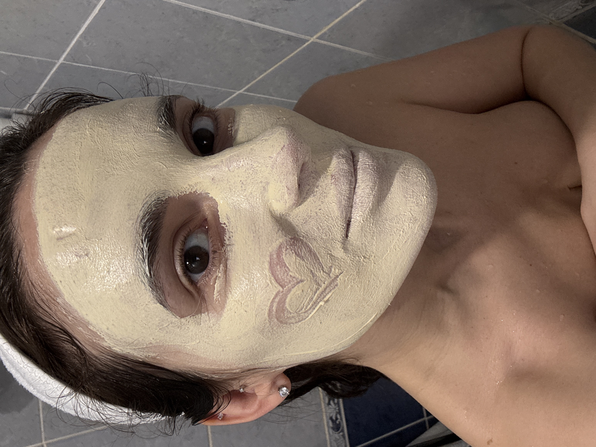 A low-angle shot depicts a woman with a face mask applied in a tiled bathroom, possibly in the city of Chaikovsky, Russia, due to the prompt's instruction. The woman's face is covered in a light-yellow mask, with a lighter pink design on the right cheek. She has dark hair pulled back with a white headband. Her eyes are open and appear to be looking directly at the camera. Her right arm is across her chest. Water droplets are visible on her chest and neck. The background consists of blue and gray tiles. The image has been rotated slightly.