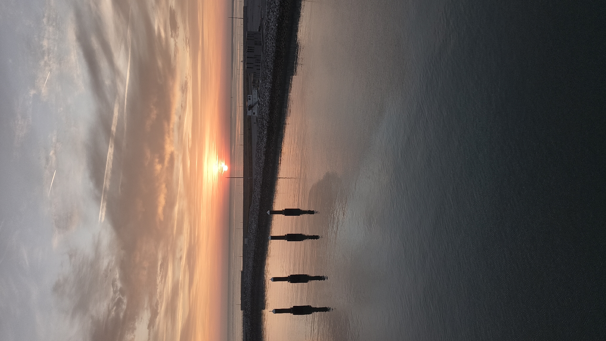 This is a vertical, medium-shot photograph of a tranquil harbor in Calais, France, at sunrise. The sun, a vibrant orange orb, is low on the horizon, casting a warm glow across the sky and reflecting on the calm water. The sky is a canvas of soft oranges, pinks, and grays, with wispy clouds streaked by faint contrails from passing aircraft.
In the foreground, the dark water is still, mirroring the muted colors of the sky. Four dark, cylindrical bollards rise from the water in a diagonal line from left to right, their reflections elongated and distorted on the surface.
Beyond the bollards, a stone-lined breakwater separates the harbor from the open sea. Along the breakwater, there are several low-rise structures, possibly utility buildings or small warehouses, their concrete facades appearing stark against the colorful sky. A few poles, likely for lighting or navigation, are visible along the breakwater and the distant shoreline. The sea is calm and appears as a muted gray-blue expanse stretching towards the horizon. The overall atmosphere is one of serene beauty and quietude, capturing the peaceful early morning in a French port city.