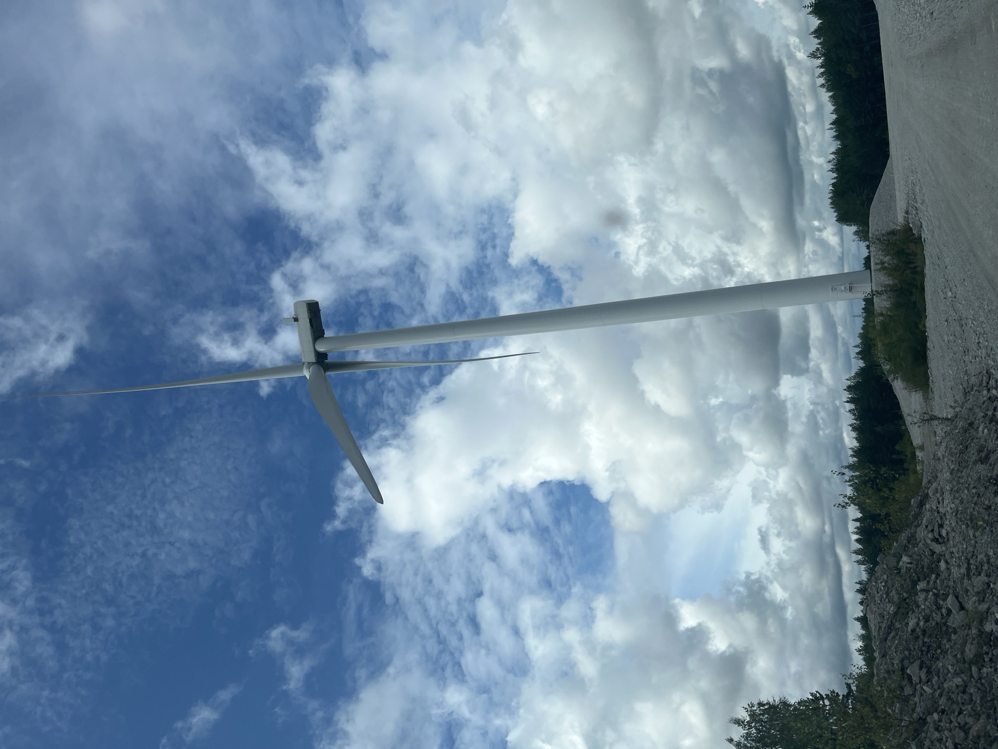 The image shows a tall, white wind turbine with three blades, one of which is clearly visible in the foreground. The turbine is located in Aidu, Estonia, on a partly cloudy day with a blue sky. The ground around the turbine is a mixture of gravel and dirt, with piles of rocks and a dirt road visible. A dense forest of pine trees lines the horizon behind the turbine. The overall scene suggests a rural or industrial landscape dedicated to renewable energy production.
