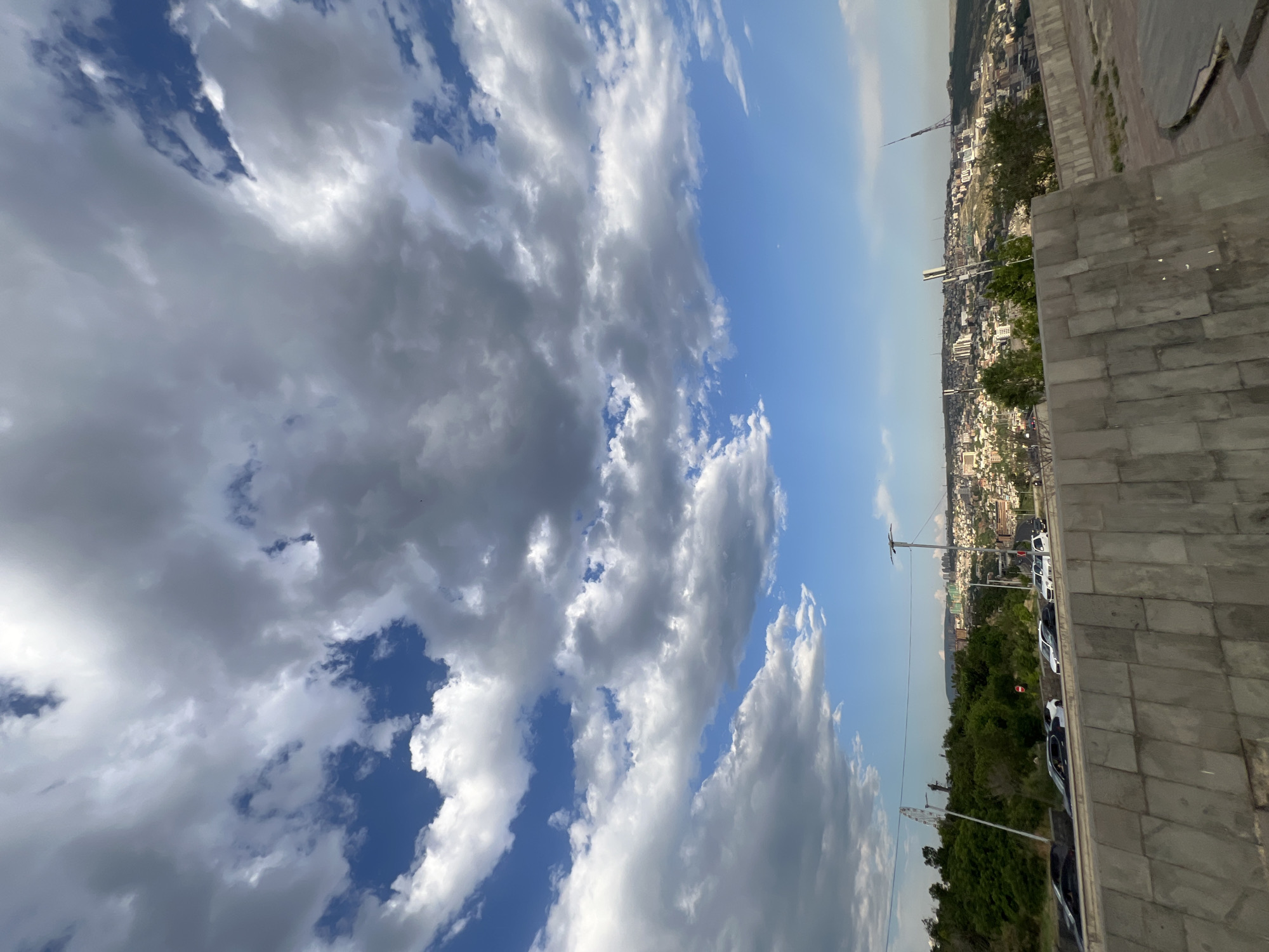 The image displays a panoramic view of the city of Yerevan, Armenia, from a vantage point that overlooks the urban landscape. In the foreground, a textured concrete wall with a rough, segmented surface runs horizontally across the bottom of the frame. Beyond the wall, a road with several cars is visible, including a dark sedan, a white sedan, and other vehicles. To the left of the road, lush green trees and foliage line a slope, interspersed with what appears to be a Ferris wheel in the distance.
The mid-ground features a sprawling city with numerous buildings, ranging in size and color, spread across rolling hills. The urban density suggests a significant population. Tall, slender structures, possibly communication towers or antenna masts, are scattered throughout the cityscape. In the far background, the landscape fades into a hazy horizon.
The sky above is dramatic, filled with a mix of bright blue patches and large, voluminous clouds. Some clouds are white and puffy, indicative of fair weather, while others are darker and more ominous, suggesting potential for rain or changing weather conditions. The lighting indicates it is daytime, likely late afternoon or early evening, given the angle of the sun and the shadows cast. There are no discernible people in the image. The overall scene captures the dynamic interplay between the natural elements of the sky and the urban environment of Yerevan.