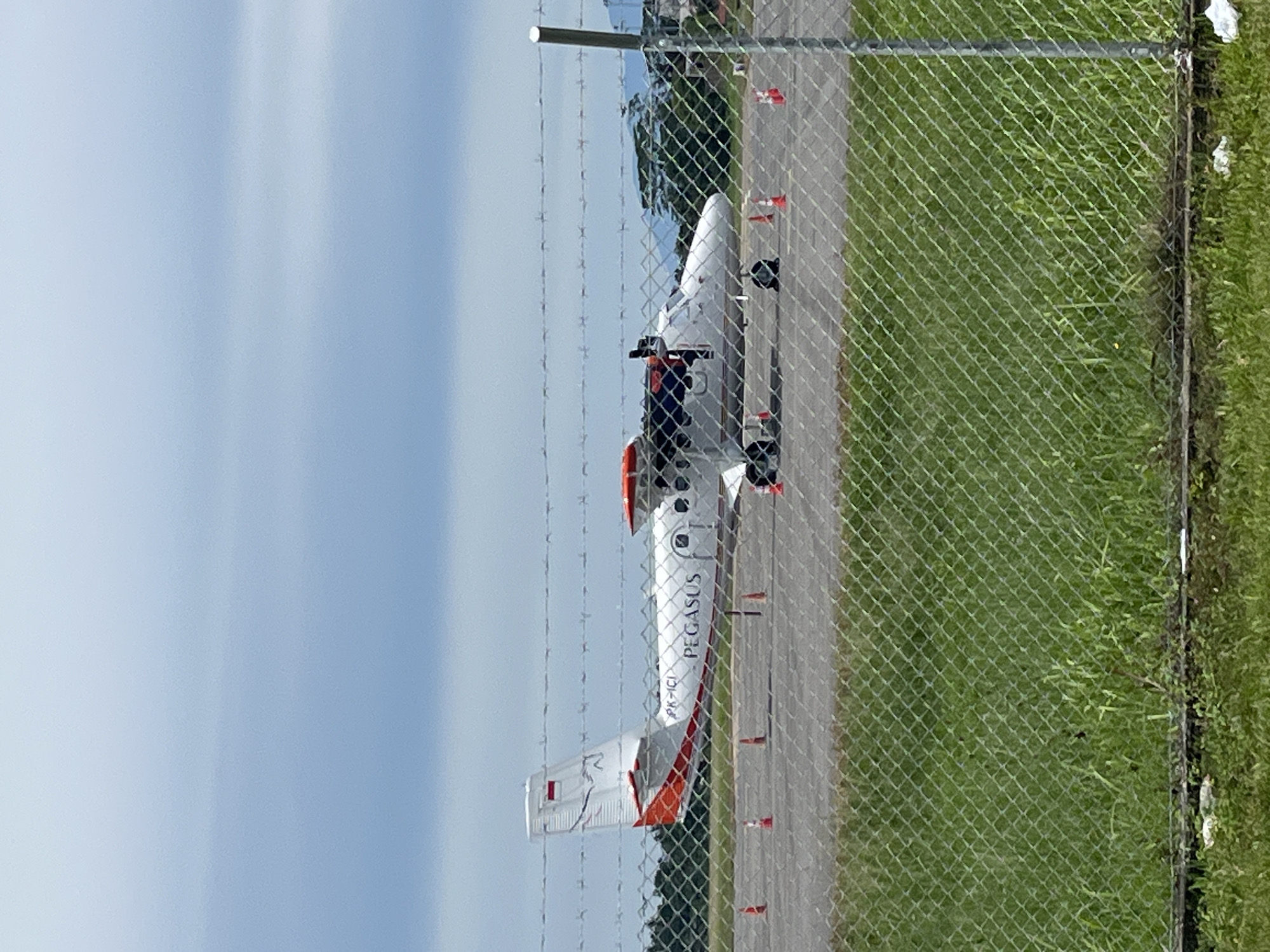 A twin-engine propeller aircraft with the registration PK-ICI and the word "PEGASUS" written on its fuselage is parked on an asphalt tarmac, as seen through a chain-link fence. The aircraft is white with orange accents on its tail and wings. The tail fin displays the Indonesian flag. Several orange traffic cones are positioned around the aircraft. The scene is set against a backdrop of green grass, trees, and a cloudy sky. This image was taken in Peurupok, Indonesia.