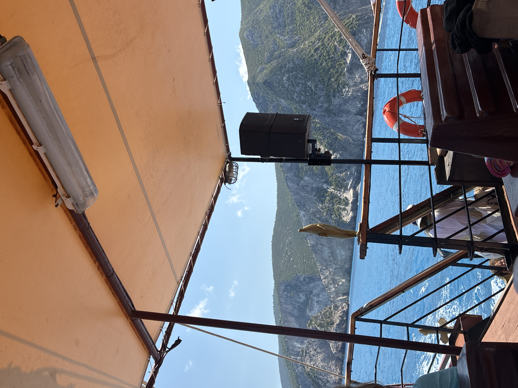 The image shows a view from the covered deck of a boat sailing on blue water. In the foreground, there are wooden seats and railings, along with orange life preservers. A black speaker is mounted on a pole. In the background, rugged, green-covered cliffs rise from the sea under a bright blue sky with some clouds. The location is the coastline near Elaties Municipal Unit, Greece.