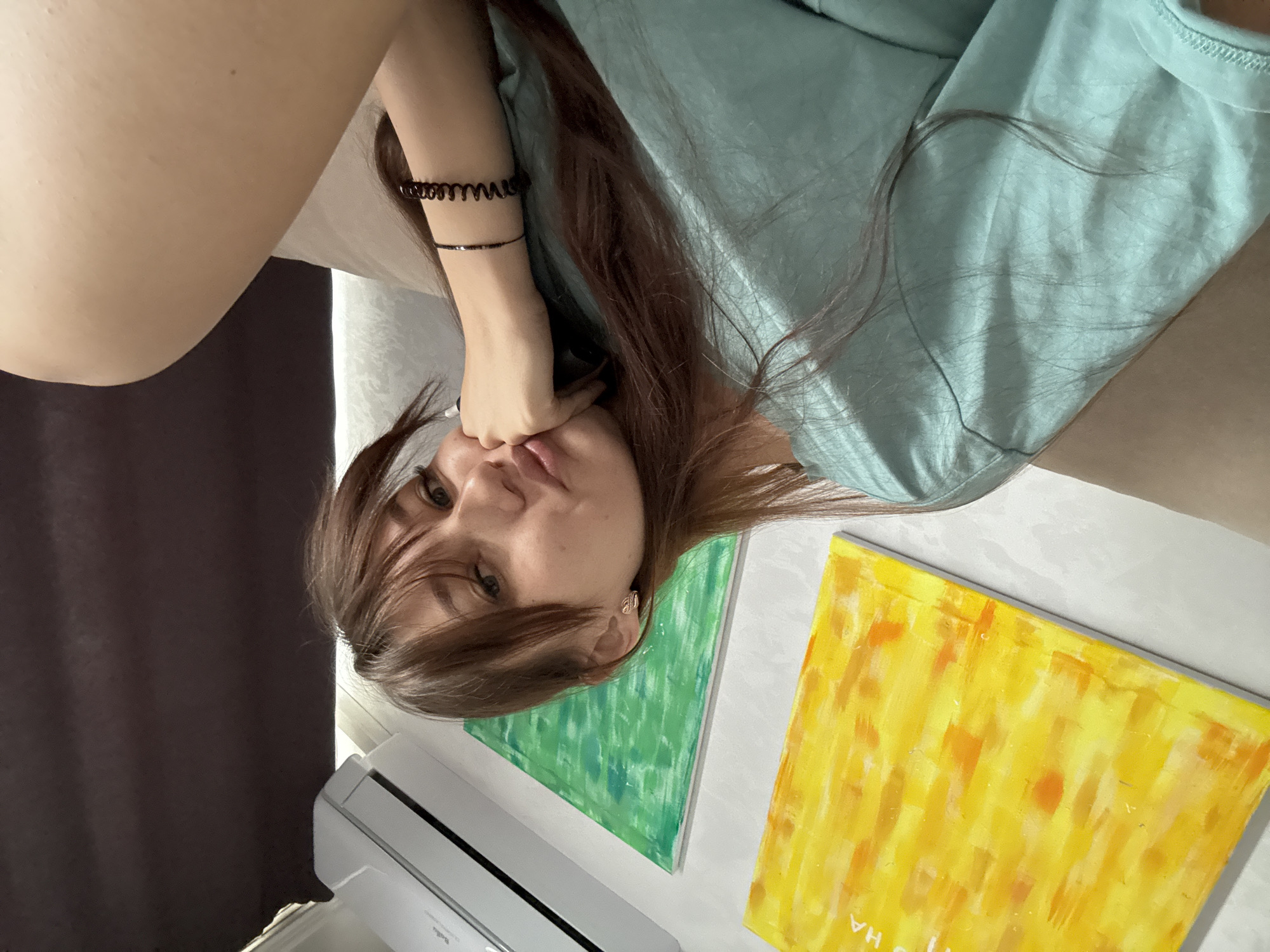 A young Caucasian woman with long brown hair and bangs sits on a couch in a living room, resting her chin on her hand. She is wearing a light blue t-shirt and has two bracelets on her left wrist. The room features two abstract paintings on the wall, one yellow and one green, and an air conditioning unit above them. A dark curtain is visible on the right side of the frame. The woman's expression is pensive. The scene is consistent with a domestic interior in Novokuznetsk, Russia.