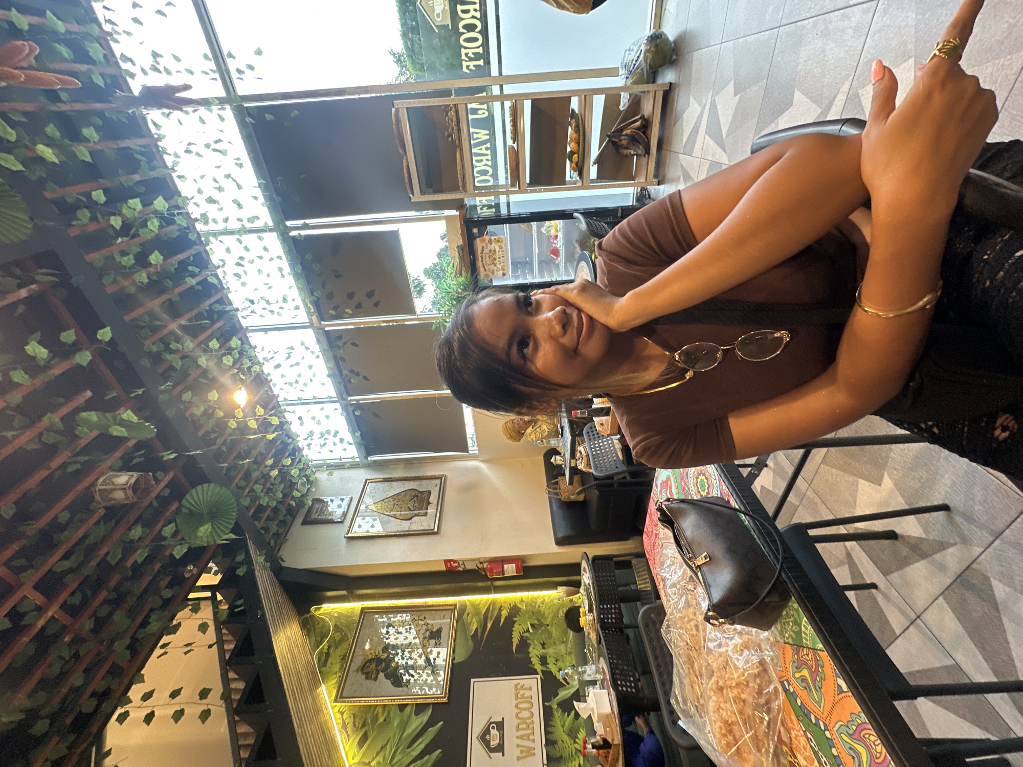 A woman with dark hair and a brown top is sitting at a table in a brightly lit cafe or restaurant. She is smiling and has one hand on her face and the other raised with a ring visible on her finger. The cafe is decorated with artificial vines and greenery on the ceiling and walls. There are framed pictures on the wall and the cafe's name, WARCOFF, is visible. The location appears to be in Central Jakarta, Indonesia. A black purse is on the table. Other tables and chairs are visible in the background.