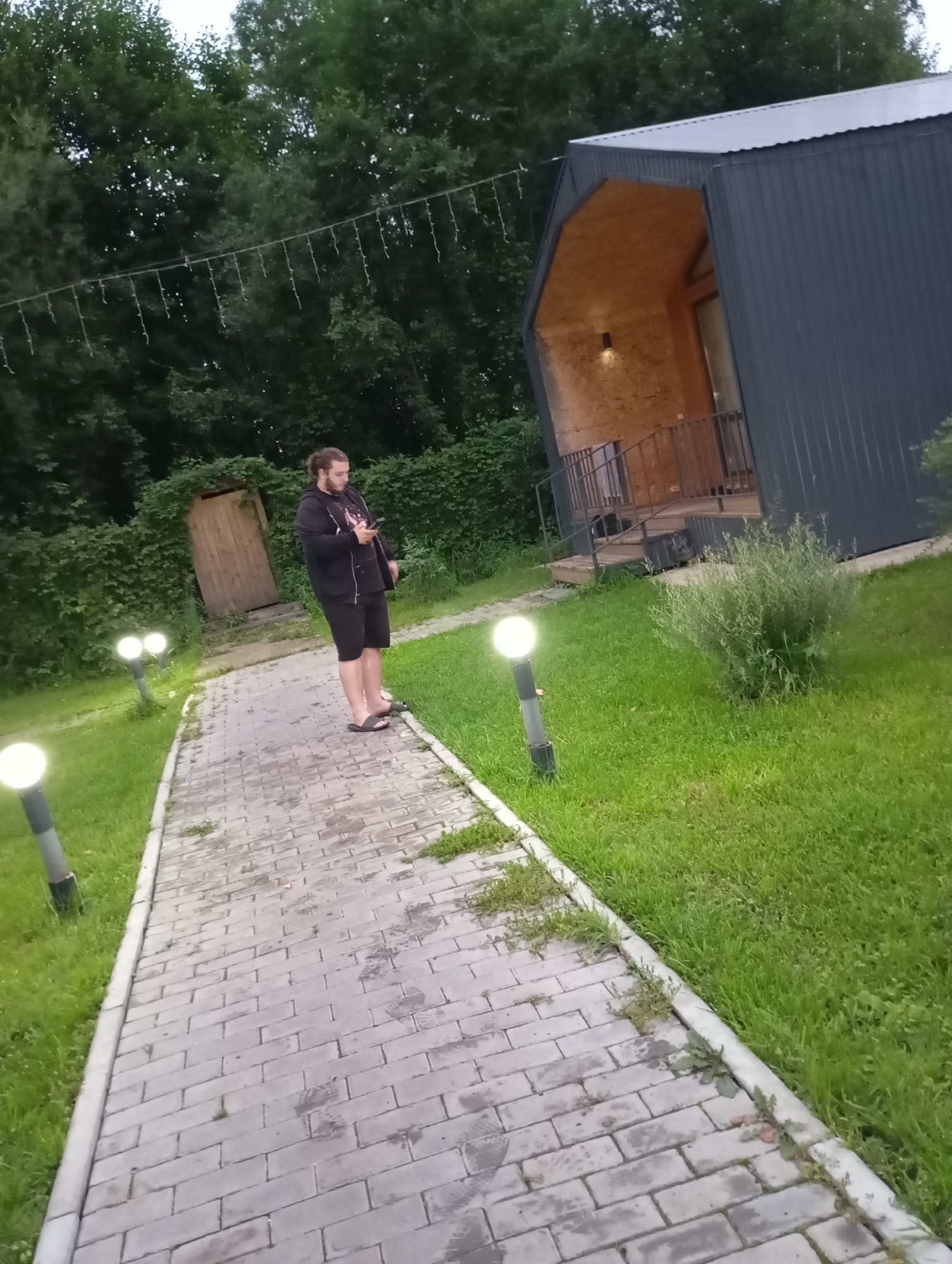 A man with light brown hair and a beard stands on a paved walkway, looking at his phone. He is wearing a black hooded jacket and black shorts, with black flip-flops on his feet. The walkway is bordered by green grass and lined with modern, spherical outdoor lamps that are illuminated. In the background, there is a wooden door set into a green hedge. Further in the distance, a modern, dark-colored structure with a slanted roof and a wooden interior is visible. The environment suggests an outdoor recreational area, possibly a park or resort, with lush greenery and a contemporary architectural design. The location is identified as Malyi Bukor, Russia.