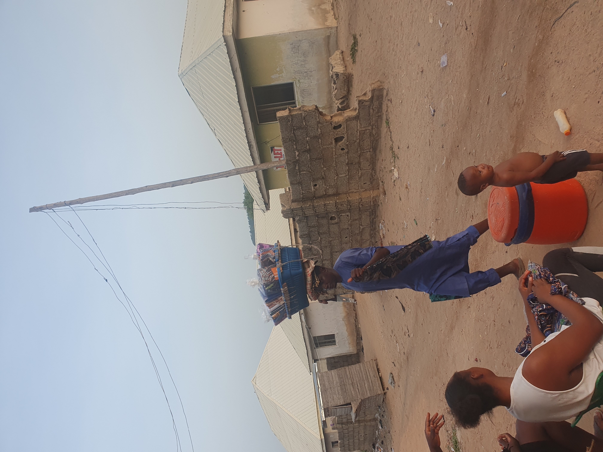 The image shows a street scene in Lokoja, Nigeria. The ground is sandy with sparse vegetation. Several buildings with corrugated metal roofs are visible. A man wearing a blue shirt and pants carries a large load balanced on his head. A young, shirtless boy stands near an orange bucket. Two women are sitting on the ground, one wearing a white tank top and black leggings and the other wearing a dark shirt. Power lines stretch across the sky.