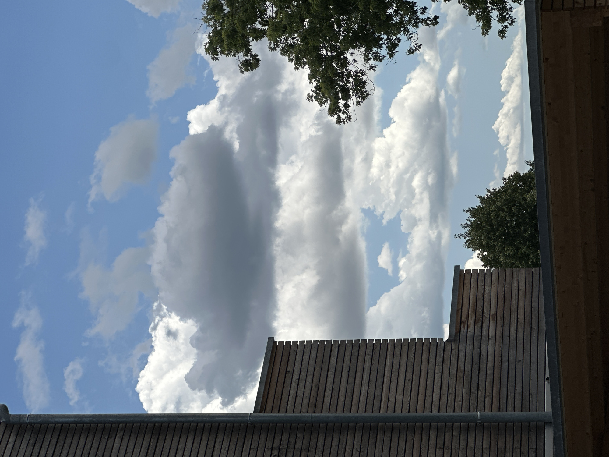 This is an outdoor, daytime, eye-level shot of a cloudy sky above a building and trees. The building has a wooden facade and a gutter pipe on the left side. In the foreground, the roof of another building is visible. To the right, a tree with green leaves is partially obscuring the sky. The sky is a mix of blue with scattered white and gray clouds. The scene appears to be in Günzburg, Germany, based on the context provided. No people or actions are depicted.