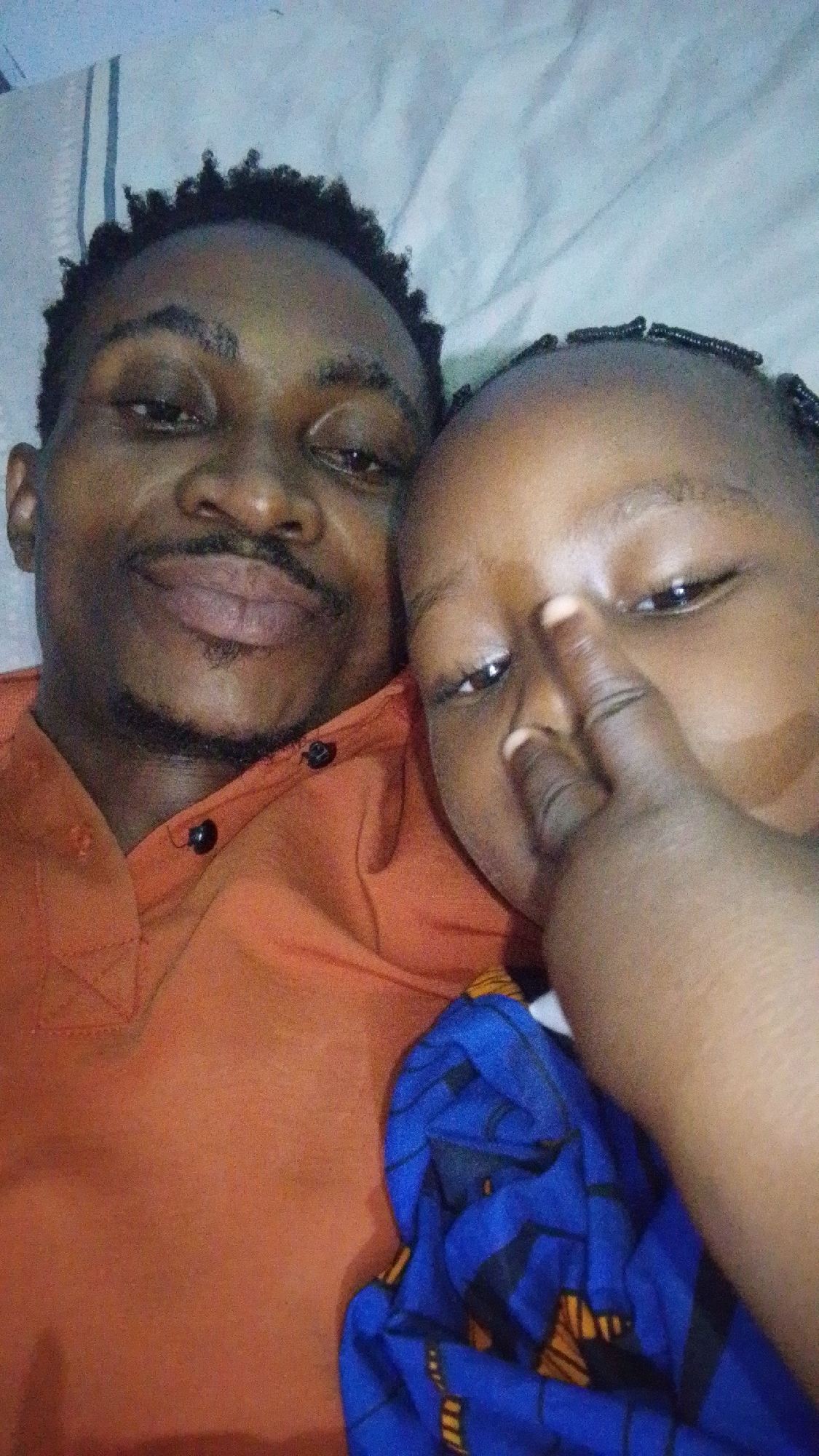 In Adankolo, Nigeria, a close-up, selfie-style photograph captures two individuals, presumably an adult and a child, lying down. The adult, positioned on the left, is wearing an orange collared shirt. The child, on the right, is partially obscured, with their finger touching their nose. A blue and yellow patterned fabric is visible near the child. The background consists of a light-colored, creased surface, possibly a bedsheet or mattress. The lighting is dim, suggesting an indoor setting, likely at night.
