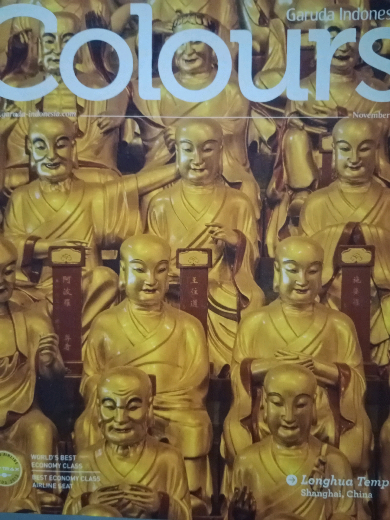The image shows a close-up of a magazine cover featuring a large font that reads "COLOURS." The background is filled with numerous golden statues of Buddhas, some with serene expressions and others with more animated faces, arranged in multiple rows. There are also some vertical banners with Asian script. In the lower left corner, text indicates "WORLD'S BEST ECONOMY CLASS" and "BEST ECONOMY CLASS AIRLINE SEAT." In the lower right corner, text reads "Longhua Temp Shanghai, China." The cover also includes the Garuda Indonesia logo and the text "November." The overall impression is that of a travel or lifestyle magazine. There is no indication of the city of Yogyakarta, Indonesia in this image.