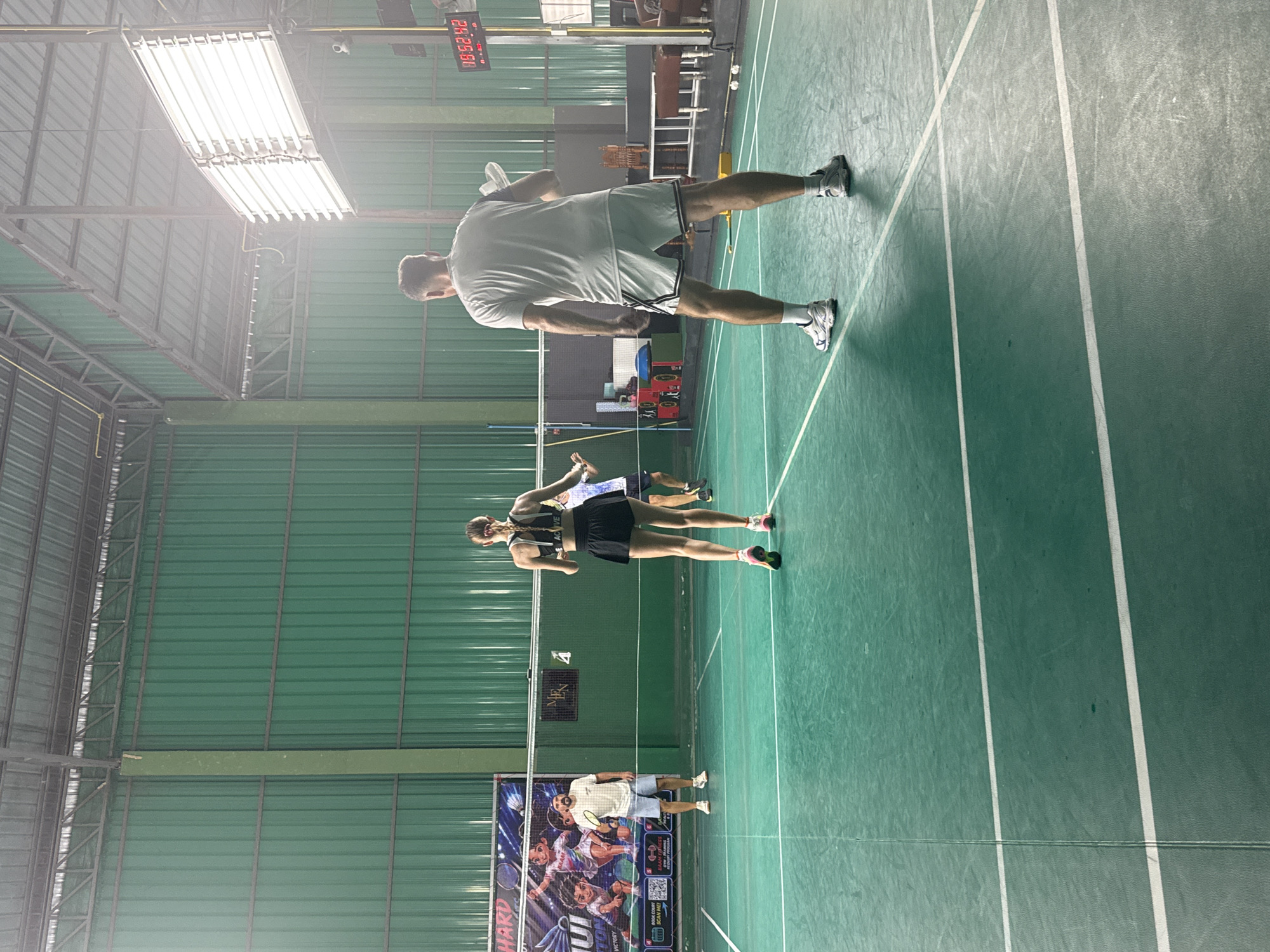 This media file shows an indoor badminton court with several people engaged in a game. The setting is an indoor sports facility with a green court marked with white lines, a high ceiling with industrial lighting, and green metal walls.
In the foreground, two individuals, a man and a woman, are playing badminton. The man, wearing a white t-shirt and light blue shorts, is holding a badminton racket and appears to be mid-swing. The woman, in a black sports bra and black shorts, is also on the court, seemingly in position to play. Further back, another man, dressed in a light grey t-shirt and grey shorts, is also present on the court, possibly observing or waiting for his turn.
In the background, a large digital scoreboard displays the time "10:52:42" in red digits. There are also posters on the wall depicting athletes playing badminton. The overall lighting suggests it is daytime, or the facility is well-lit, as evident from the bright ceiling lights. The atmosphere appears active and competitive, typical of a sports game.
Visible text includes the time on the scoreboard, and potentially text on the posters in the background, though it is not clearly legible. The context of Ko Samui District, Thailand, implies a tropical climate, but this is not directly evident from the image as it is an indoor scene.