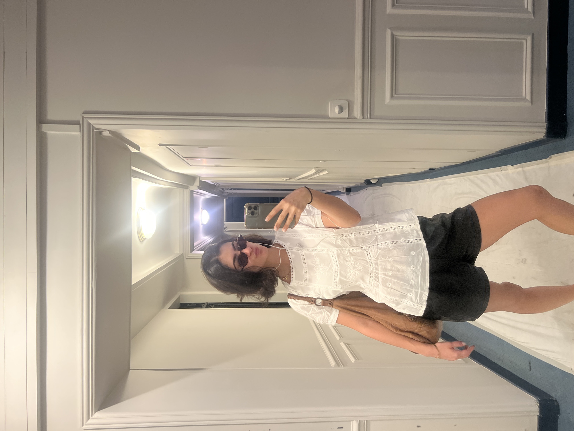 A young woman with dark hair, wearing sunglasses, a white blouse, and black shorts, poses for a selfie in what appears to be a hotel room. She holds her phone in her right hand, facing a mirror. The room is clean and sparsely furnished, with a white bed and blue carpet along the wall. The door to the room is visible, as is a glimpse of a hallway with lighting. The room's design suggests a standard hotel. The location cannot be determined to be in Paris, France based on visual data alone.