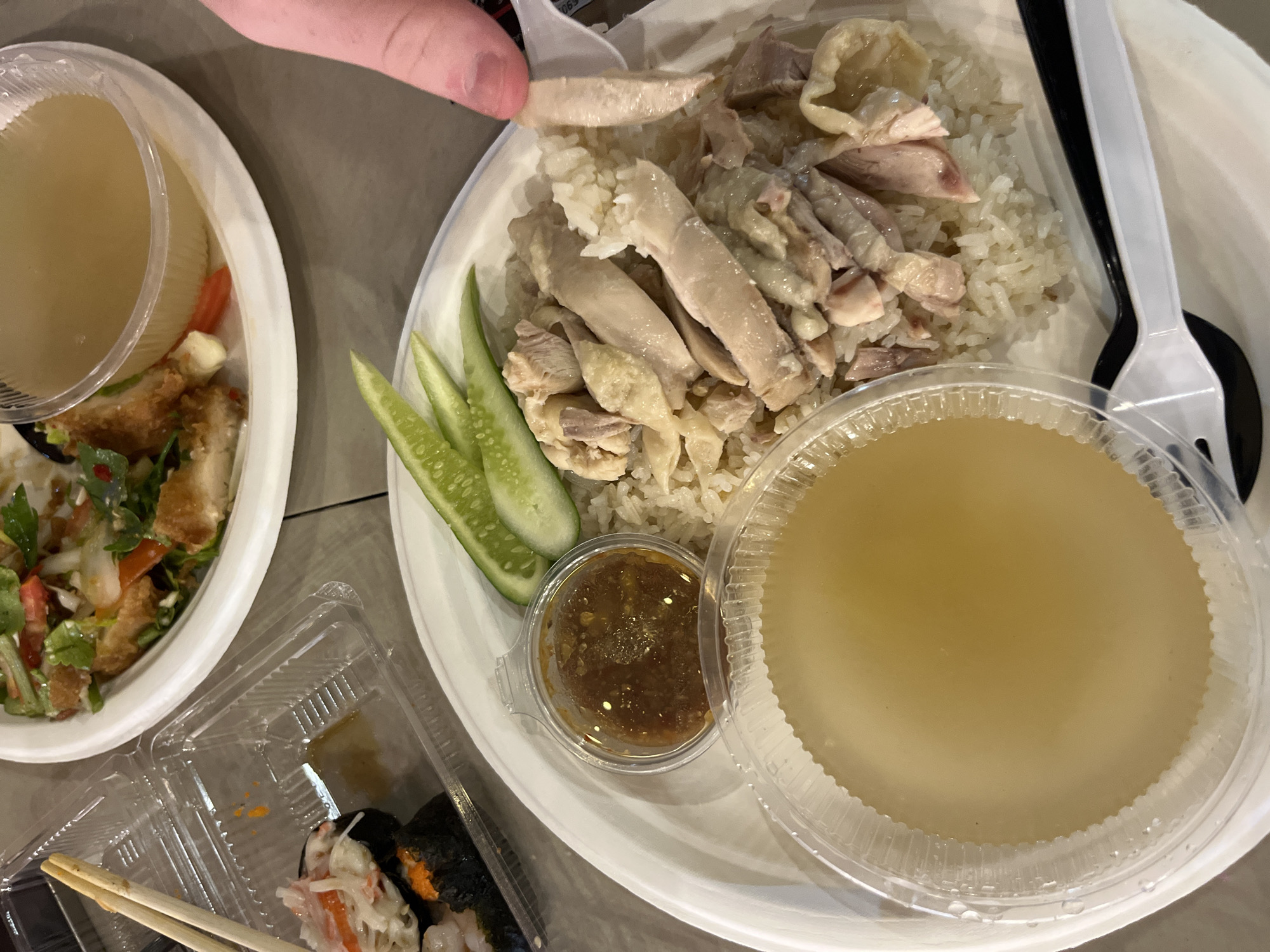 The image shows a meal consisting of Hainanese chicken rice, salad, and sushi, arranged on a gray table. The Hainanese chicken rice is on a white plate with sliced chicken, rice, cucumber slices, a small container of sauce, and a container of broth. A white fork and a black spoon are positioned on the plate. A hand is holding a piece of chicken with a white utensil. To the left is a separate white plate containing a salad. There is also a clear plastic container with sushi. The location is Pattaya City, Thailand.