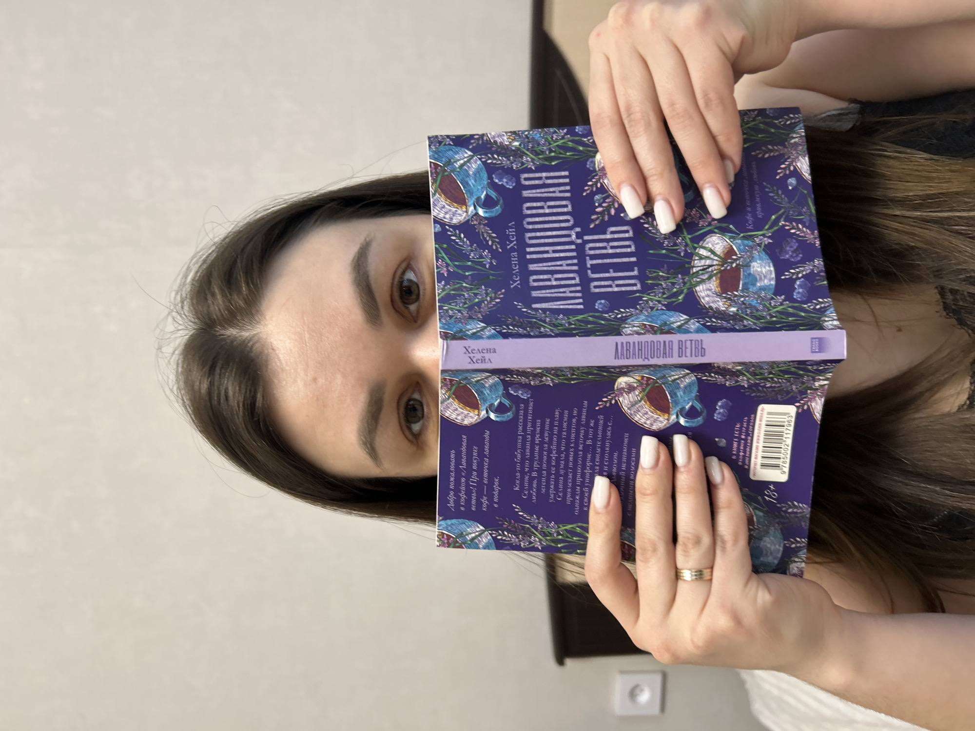A woman is holding a book up to her face, partially obscuring it. The book is titled "Lavandovaya Vetv" ("Lavender Branch") by Helena Hale, written in Russian. The book cover is purple and features illustrations of teacups and lavender sprigs. The woman has long brown hair, fair skin, and is wearing white nail polish. She is wearing a ring on her ring finger. The background is a plain, light-colored wall with a white electrical outlet. The location is Chaikovsky, Russia.