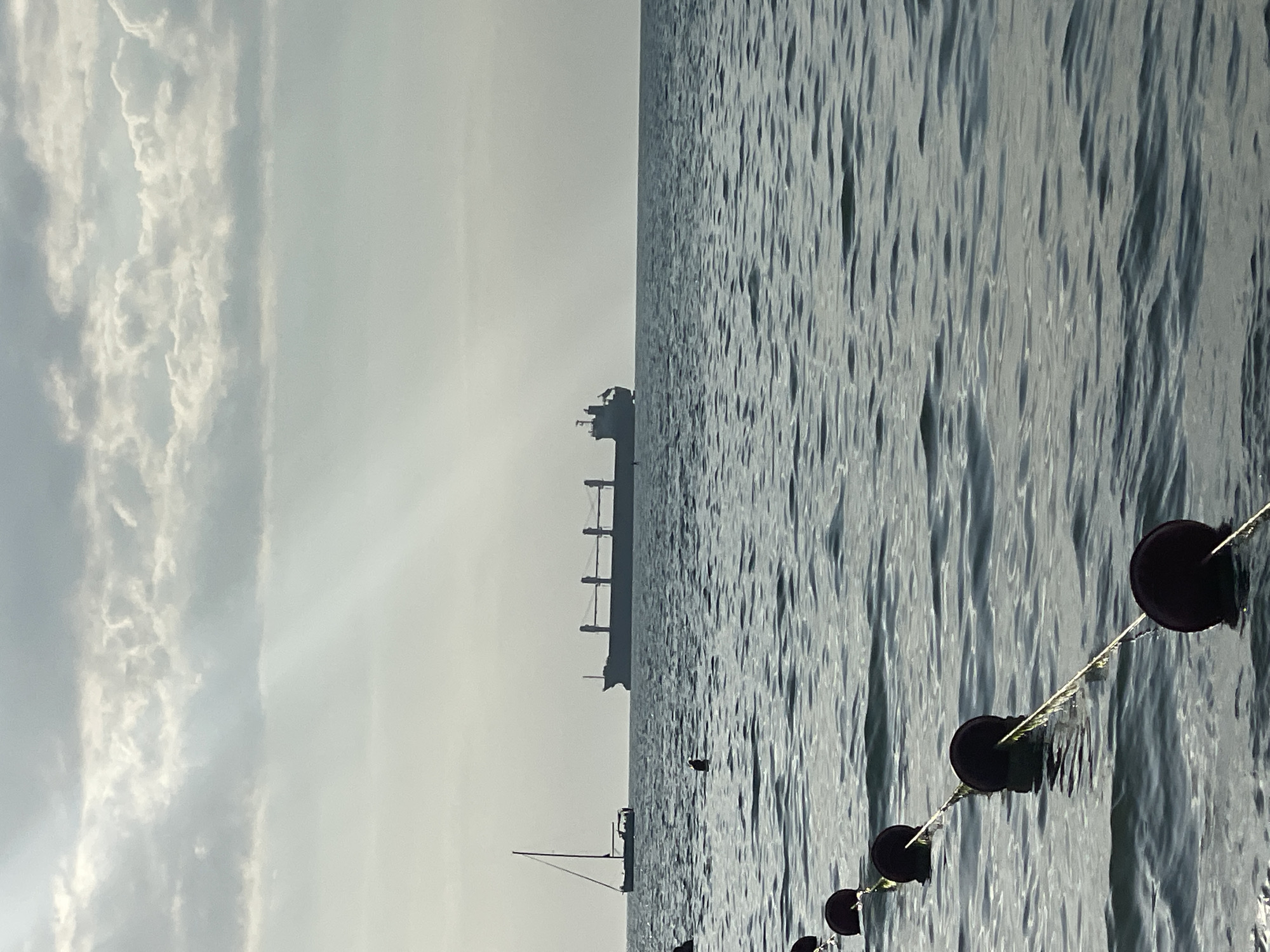 The image depicts a calm sea and sky with a large cargo ship silhouetted on the horizon. The water is rippled, reflecting the overcast sky, which is filled with wispy clouds. A string of dark buoys is visible in the foreground, creating a diagonal line across the lower right portion of the frame. These buoys are spaced apart and appear to be tethered by a rope. A smaller sailboat is also visible in the distance, to the left of the cargo ship. The overall atmosphere is peaceful and serene, suggesting a quiet day at sea. The light suggests it could be morning or late afternoon, but it's difficult to tell definitively due to the overcast conditions. The location is Anapa, Russia, which is known for its Black Sea coastline. There is no visible text in the image.