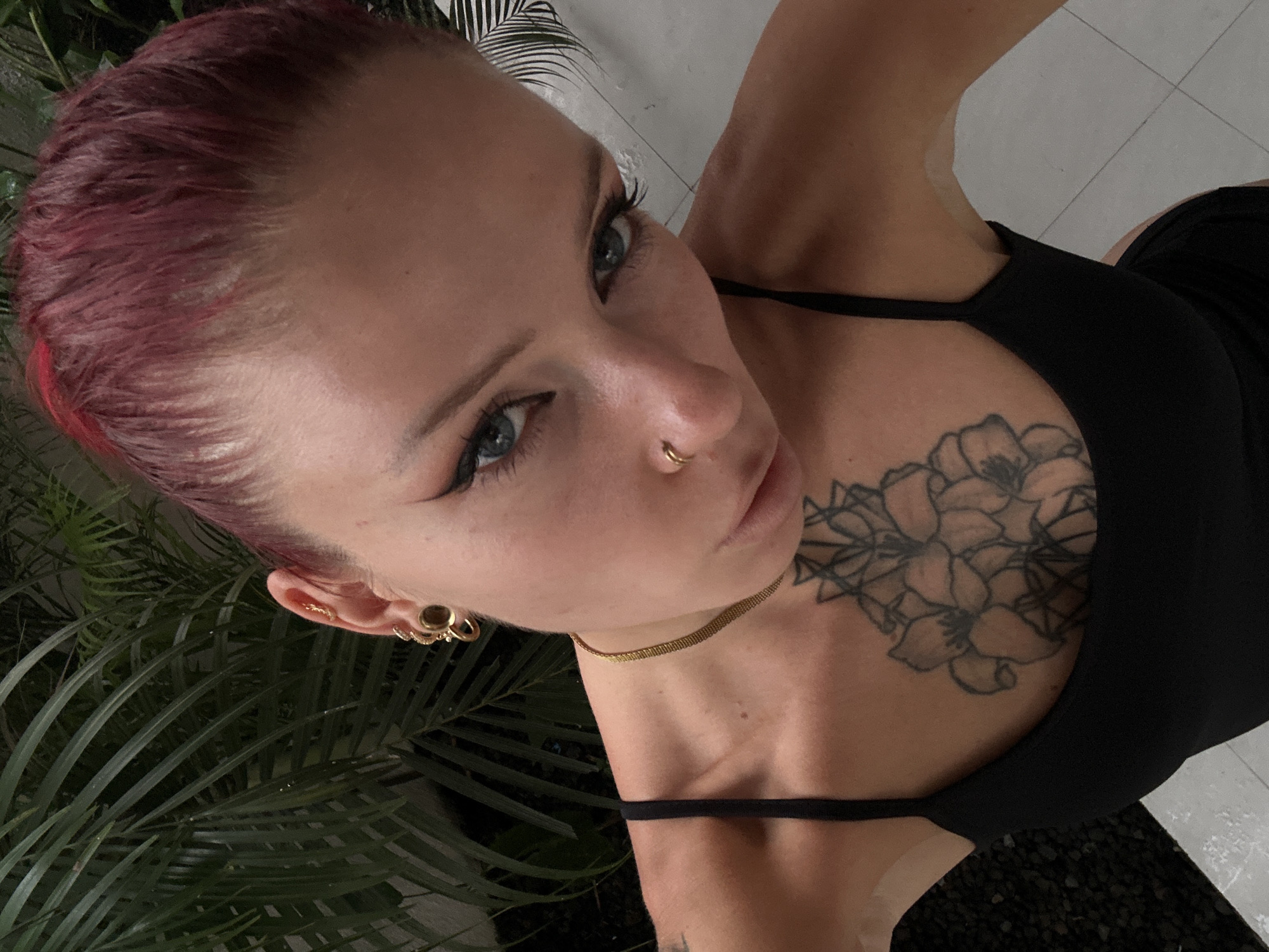 In North Kuta, Indonesia, a woman with short, reddish-pink hair, blue eyes, and a nose ring poses for a selfie. She wears a black tank top adorned with a flower tattoo on her chest. Multiple gold earrings, including hoops and studs, accent her ears, and she sports a thin gold chain necklace. The background features lush green foliage and a tiled floor, suggesting an indoor or enclosed outdoor setting.