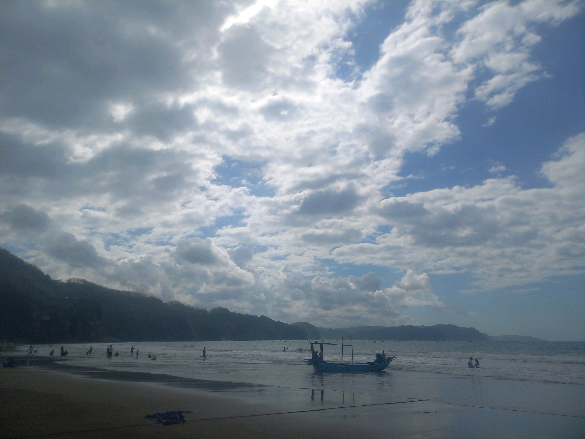 The image captures a scenic view of a beach in Indonesia. The sky is filled with a dramatic display of clouds, with patches of blue sky visible between them. A distant shoreline is marked by a line of lush green hills. The water is calm, with gentle waves lapping at the shore. Several people are scattered across the beach and in the shallow waters, some of them wading or playing. A small, blue boat is moored near the shore, with a couple of individuals visible on board. The overall atmosphere is serene and tranquil, with a hint of the vibrant life that characterizes the Indonesian coast.