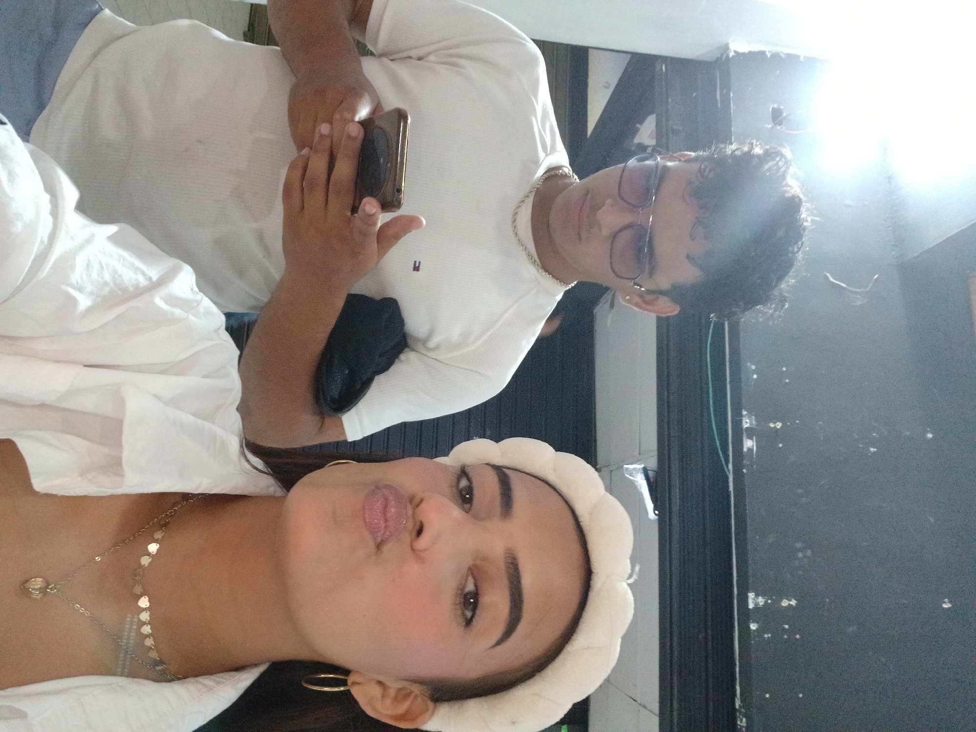 A selfie captures two individuals indoors. The woman in the foreground wears a white headband, gold hoop earrings, and a delicate gold necklace, posing with pursed lips. Behind her stands a man in a white t-shirt and glasses, holding a phone. A gold chain is visible around his neck. The background includes a door or wall. The location is not discernible as Porlamar, Venezuela.