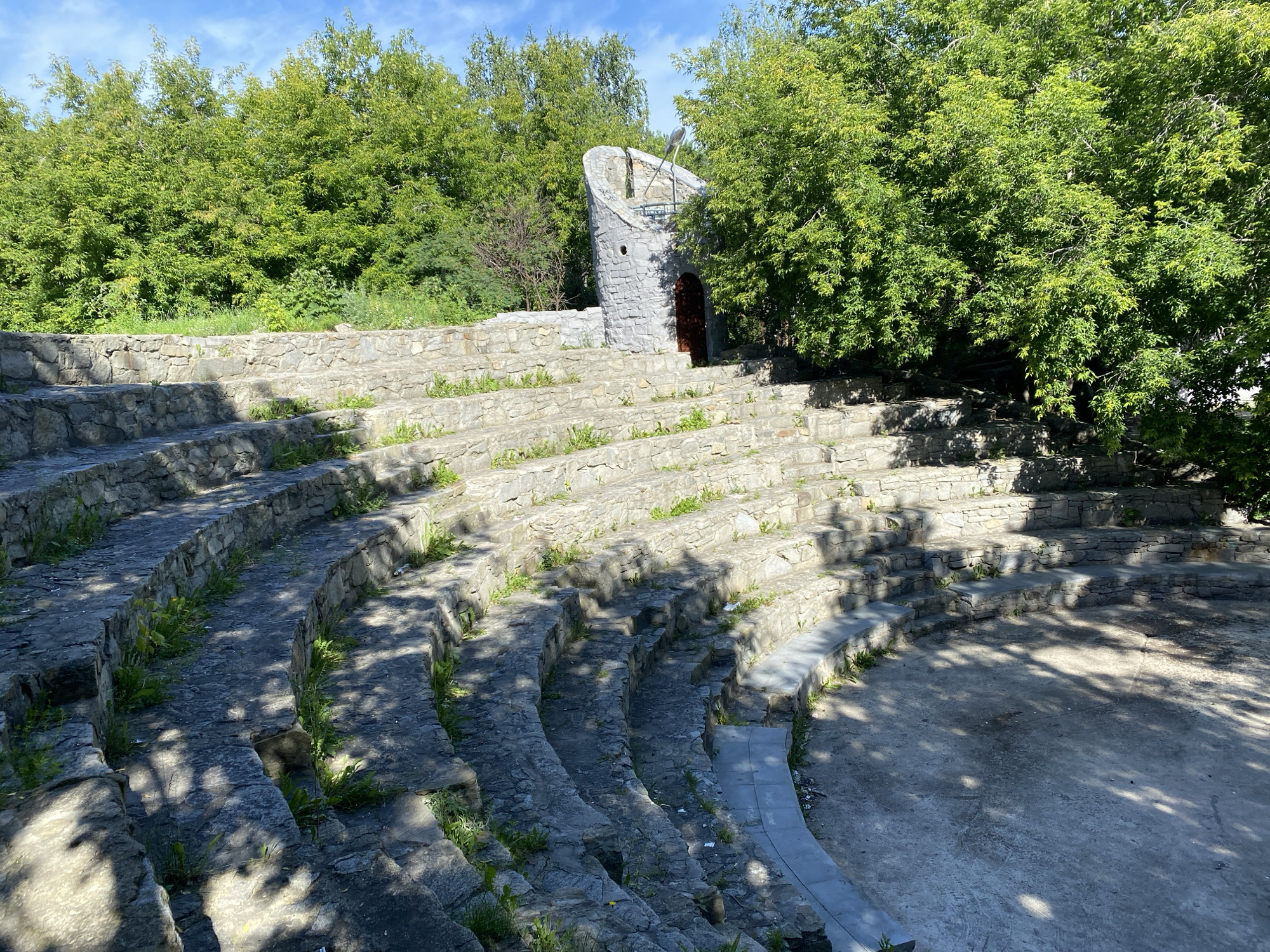 This is an outdoor amphitheater constructed from rough-hewn stones. The seating area consists of multiple curved tiers that descend towards a circular performance space. Vegetation, including grass and small plants, grows between the stones and on the edges of the seating. Behind the amphitheater, dense green trees form a backdrop, and the sky above is blue with some clouds. A small, stone tower-like structure with a arched doorway and an opening at the top stands to the right of the amphitheater. The ground in the performance area is bare earth or gravel, with shadows cast by the surrounding trees. The image appears to have been taken on a sunny day, with dappled sunlight and shadows falling across the stone seating. This location is the Stone Amphitheater in the Shartash Stone Town Park in Yekaterinburg, Russia.