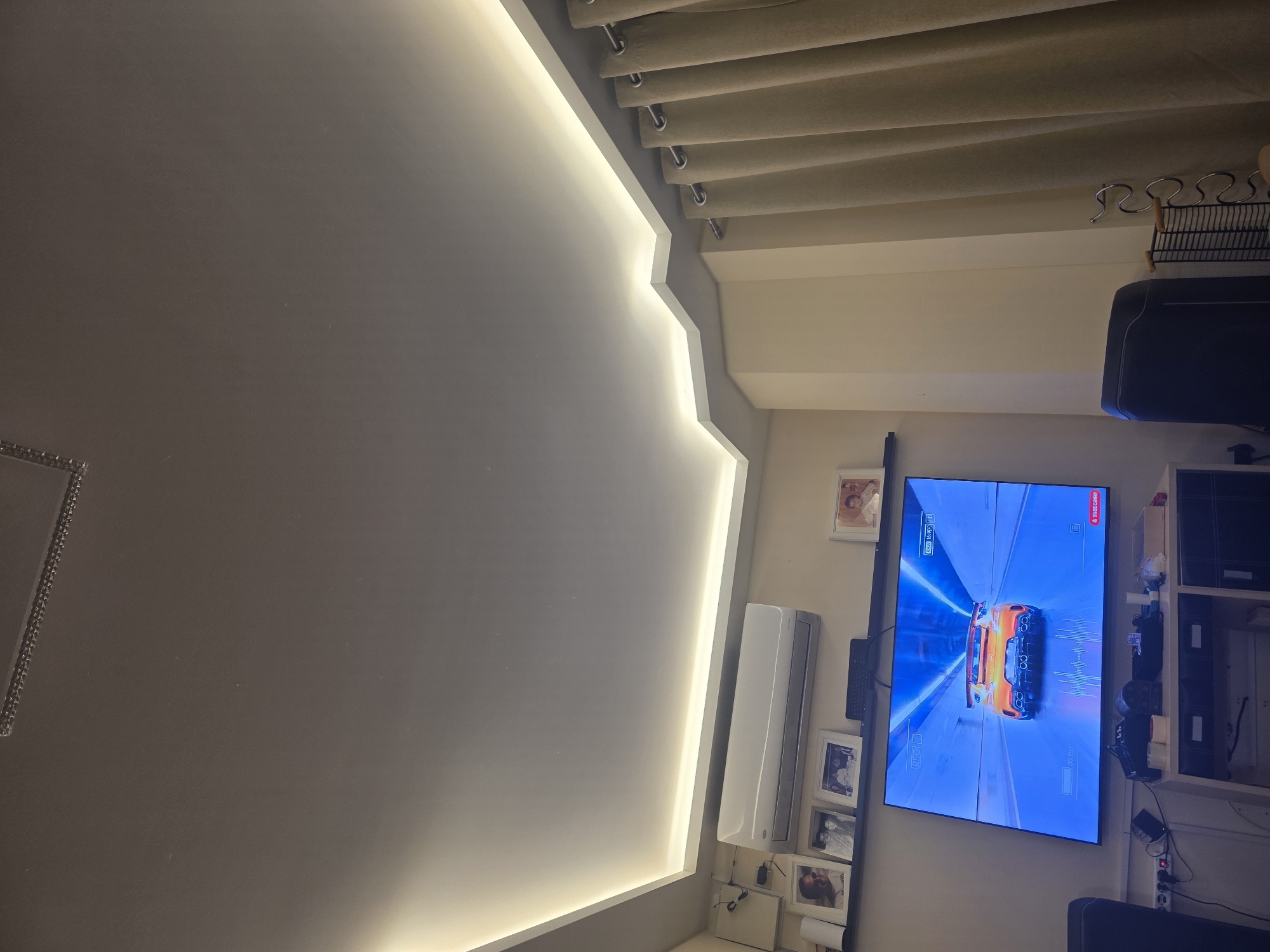 The image depicts an interior scene, likely a living room, with a focus on a large television screen displaying a car racing game. The television is mounted on a wall above a white cabinet containing drawers and shelves, with various items placed on top, including speakers, a remote, and a chest. To the left of the television, an air conditioning unit is installed on the wall, with framed photographs displayed on a shelf beneath it. The room features a modern ceiling with integrated LED lighting that casts a warm glow. On the right side of the image, beige curtains are drawn, covering a window or doorway. The overall atmosphere is dimly lit, suggesting it might be evening or the blinds are closed. There is no visible indication of the specific location within the Municipal Unit of Asini, Greece; the description is limited to the observable elements within the frame.