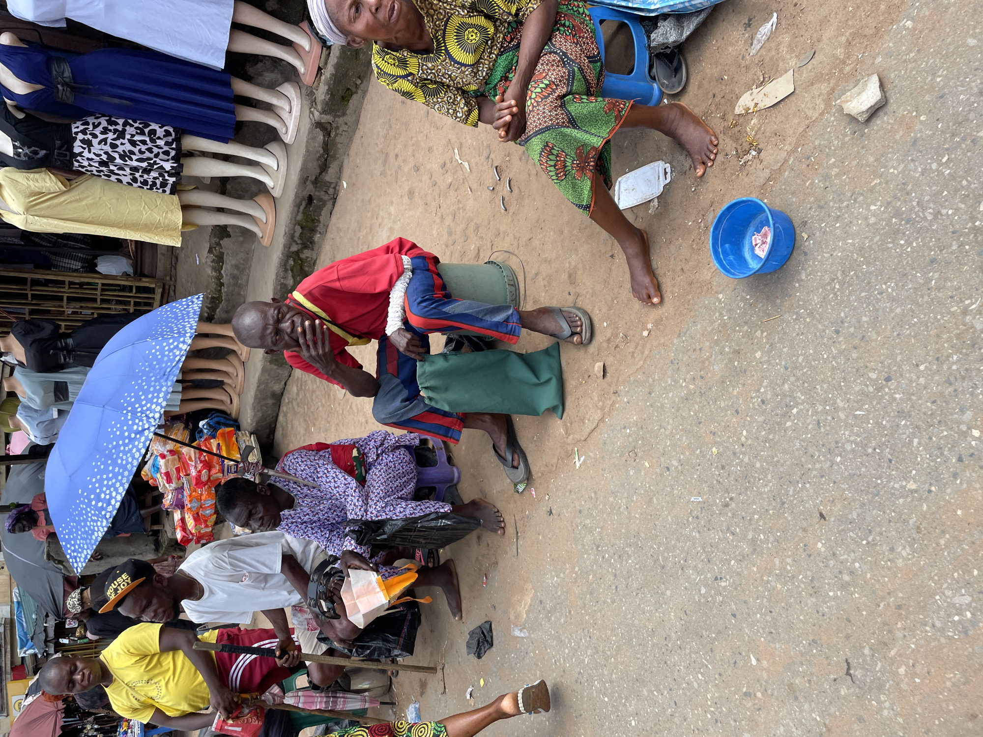 This is a medium shot, eye-level, outdoor photograph of a market scene. The foreground features a section of paved ground with scattered debris, including pieces of paper, a broken rock, and a blue plastic bowl containing a few pieces of raw meat. To the left, the bare feet of a person are visible, wearing open-toed sandals. In the midground, three men are seated on stools or buckets. The man on the far left wears a yellow t-shirt and red shorts, holding a walking stick. Beside him, a man with a black baseball cap that reads "HOUSE OF JOY" wears a white t-shirt and shorts. The third man, seated in the center, wears a purple patterned robe and is looking down. To the right of the seated men, a woman in a brightly colored yellow and green dress is seated on a blue stool, looking to her right. A large blue umbrella with white dots is partially visible behind the seated men. In the background, clothing on mannequins is displayed outside a shop. The overall scene depicts a busy street market environment.