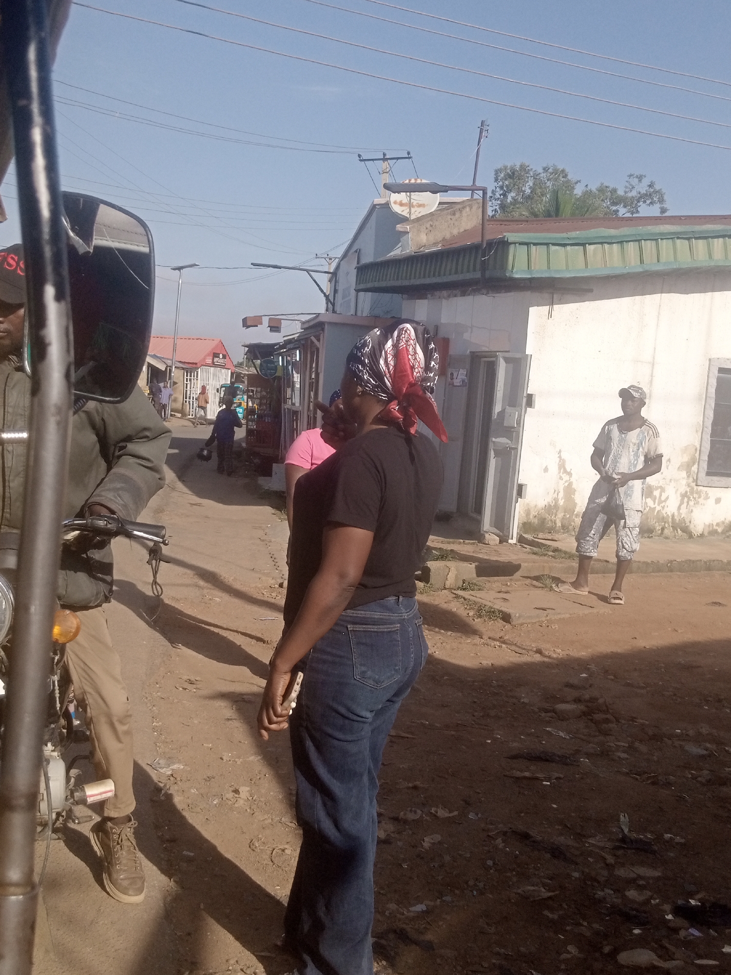 The image depicts a street scene in Gura Top, Nigeria, bathed in bright sunlight. The foreground features a person on a motorcycle, partially obscured by the vehicle's structure. Further down the dusty street, several individuals are visible. A woman in a black t-shirt and blue jeans is facing away from the camera, her head adorned with a patterned headscarf. To her right, a man wearing a light-colored patterned shirt and patterned trousers stands near a white building with an open doorway. In the background, more figures are present, and the street is lined with buildings and what appear to be market stalls. The environment is unpaved, suggesting a typical Nigerian street.