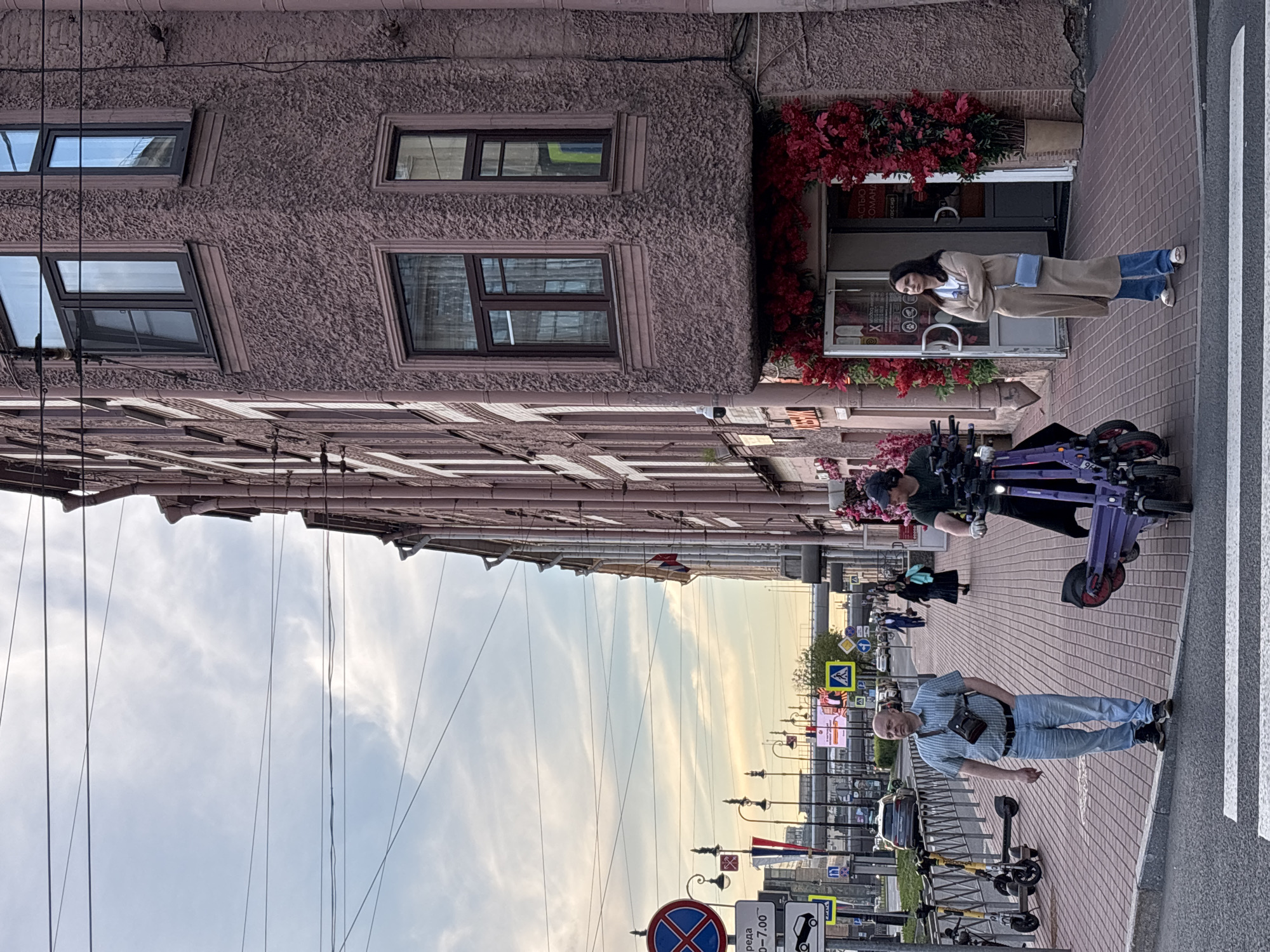 This image depicts an urban street scene in Saint Petersburg, Russia, observed during what appears to be late afternoon or early evening, characterized by the yellowish glow in the cloudy sky. The perspective is from the sidewalk, looking down a street lined with buildings.
In the foreground, on a reddish-brown brick sidewalk, a bald man wearing a light blue plaid shirt, jeans, and a black shoulder bag stands near a white-striped crosswalk. To his left, several yellow electric scooters are parked. Further down the sidewalk, closer to a building, another person in a dark cap is partially visible beside a purple electric scooter or moped.
The right side of the frame is dominated by a tall, reddish-brown building with a textured facade and dark-framed windows. At street level, a shop entrance is elaborately decorated with vibrant artificial red flowers and foliage. A woman in a long beige coat, blue jeans, and a light blue shoulder bag stands in front of this shop, looking casually to her right. Text visible on the shop door includes fragments like "АСТЬЮ", "ОМАНА", and "КАССА" (CASHIER).
The left side of the street features a long row of buildings, stretching into the distance. Numerous lampposts and various traffic signs are visible, including a red circle with a blue 'X' (No Stopping/Standing) and a yellow square with a pedestrian symbol (Pedestrian Crossing). Some Cyrillic text, such as "среда 9:00-7:00", is also present on a white sign. Power lines crisscross the cloudy sky above the street. The overall atmosphere is that of a bustling city street at dusk.