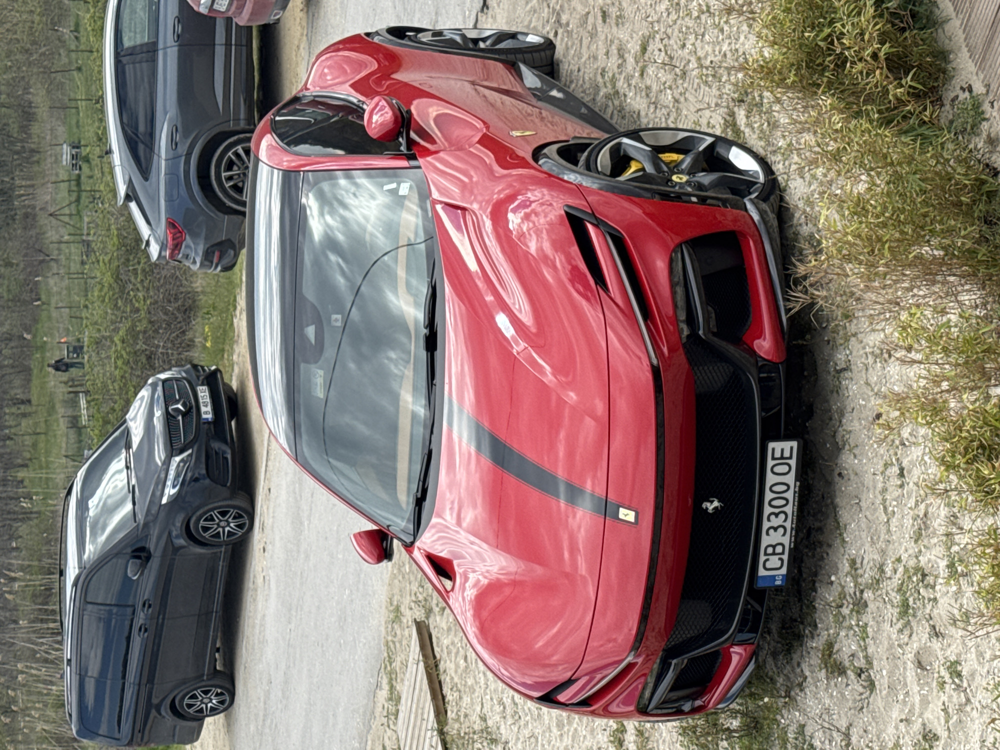 The image depicts a red Ferrari parked next to a dirt path, with two other vehicles – a black Mercedes-Benz van and a dark grey SUV – parked behind it. The Ferrari's license plate reads "CB 3300 OE," indicating it is registered in Bulgaria. The surrounding environment consists of a gravel path and some dry vegetation. The presence of Bulgarian license plates suggests the location is indeed in Bulgaria, and considering the license plate code "CB," Varna is a probable location.