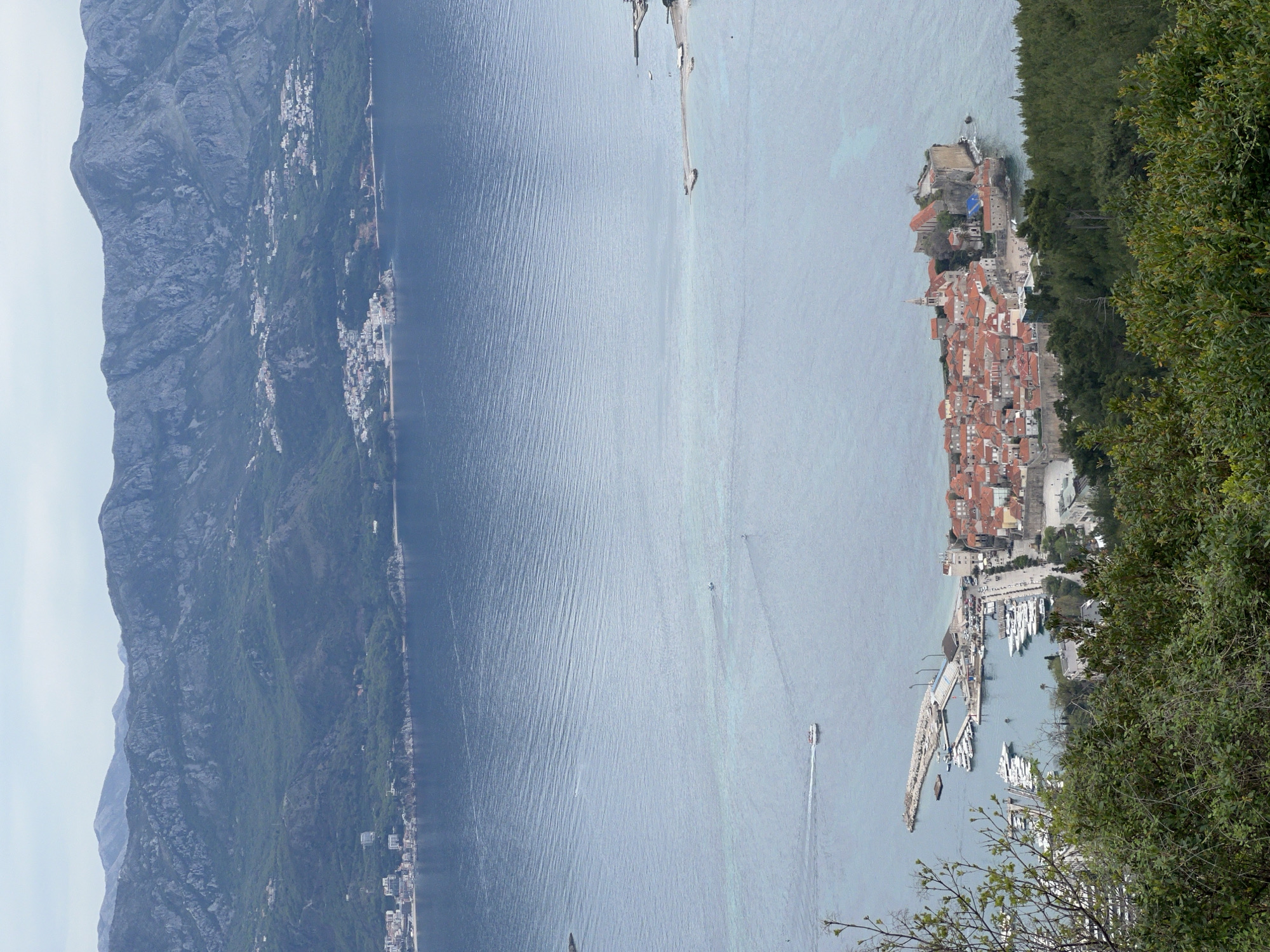 The image shows an aerial view of Sveti Stefan, a small islet and 5-star hotel resort in Montenegro, located on the Budva Riviera. The islet is connected to the mainland by a narrow isthmus. It has characteristic red-tiled roofs. The waters surrounding the peninsula are calm, with boats visible in the bay and a long pier jutting out of the mainland. High mountains are visible on the left side of the image, while dense green vegetation frames the scene on the right. The image is taken from an elevated position, likely a nearby mountain.
