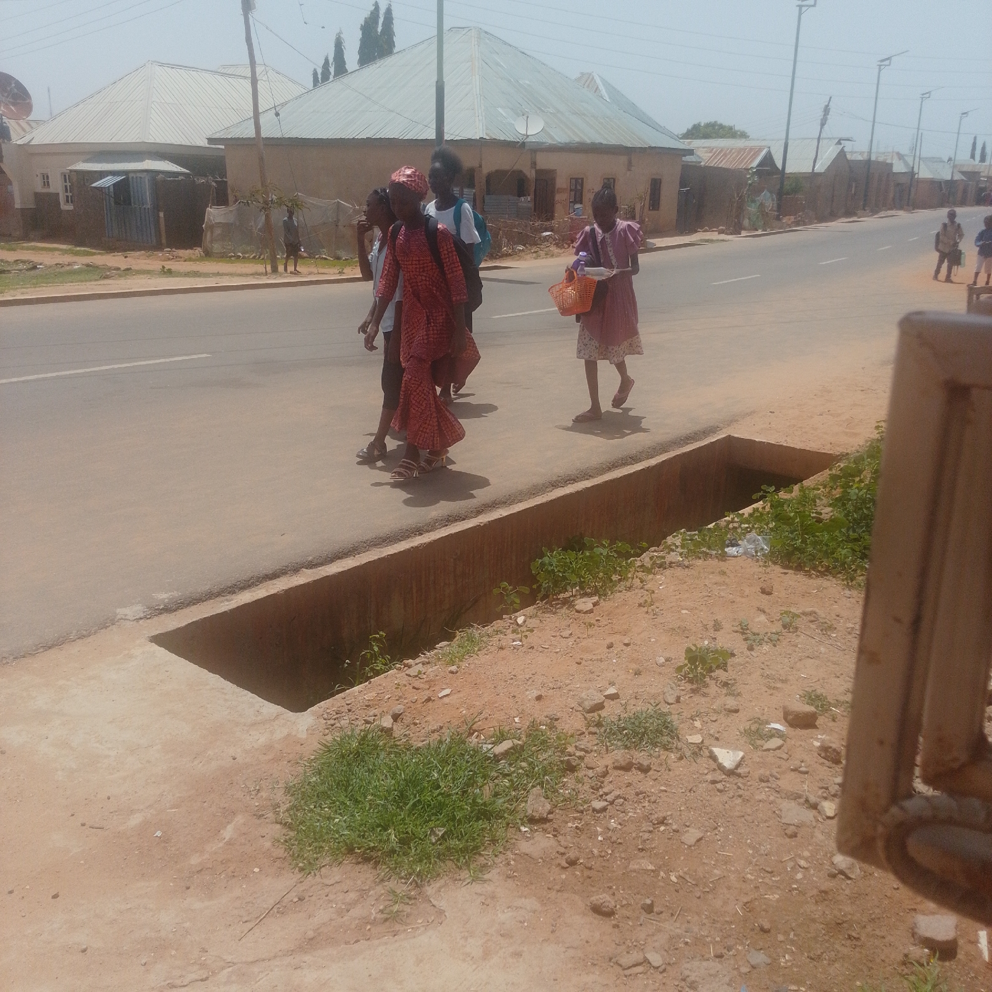 A group of four young girls walk along a paved road in Gombe, Nigeria. The girl in the foreground on the left wears a red and orange patterned dress and a matching headscarf, carrying a black backpack. To her right, another girl in a white top and black skirt also wears a black backpack. A third girl in a pink dress walks further ahead, holding an orange basket with a white container inside. The fourth girl, to the far right, is mostly obscured but appears to be wearing a patterned dress. They are walking on the side of a two-lane road with a white line marking the center. Buildings with corrugated metal roofs line the far side of the road. A concrete drainage ditch runs along the near side of the road, with sparse vegetation and some scattered rocks. The overall environment appears to be an urban or semi-urban area.