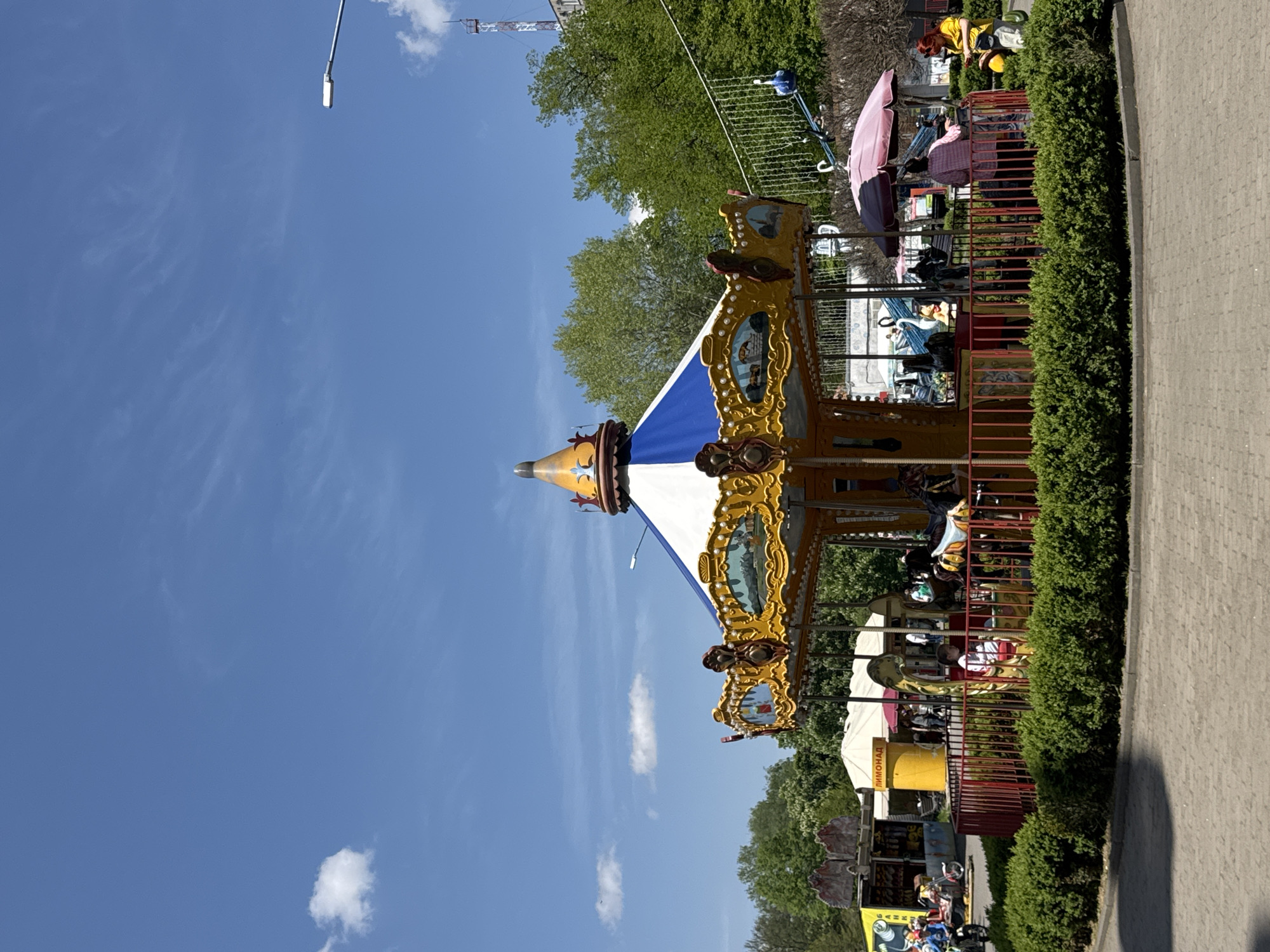 Captured in Vladikavkaz, Russia, the image showcases a classic carousel positioned beside a paved walkway, bordered by meticulously maintained hedges. The carousel features a blue and white striped conical roof, adorned with ornate gold detailing. The ride itself is enclosed by red railings. A pink parasol provides shade near the carousel.
In the background, verdant trees fill the space, framing the scene. A utility pole is seen against the clear blue sky dotted with wispy clouds. Other structures, including a broadcast tower, are partially visible through the trees.