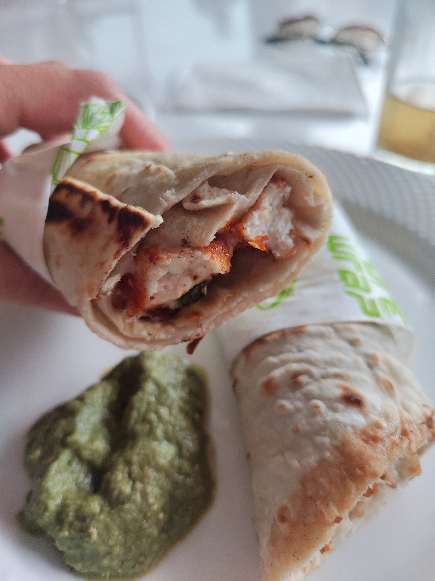 A close-up shot depicts a hand holding a partially unwrapped chicken wrap. The wrap contains cooked chicken and a sauce, encased in a flatbread. A green sauce, presumably a chutney, is present next to another wrapped roll on a white plate. In the background, a pair of eyeglasses and a glass of liquid are visible, suggesting an indoor setting. There is no definitive indication of the image being specifically located in Dharamkot, India, just based on visual information.