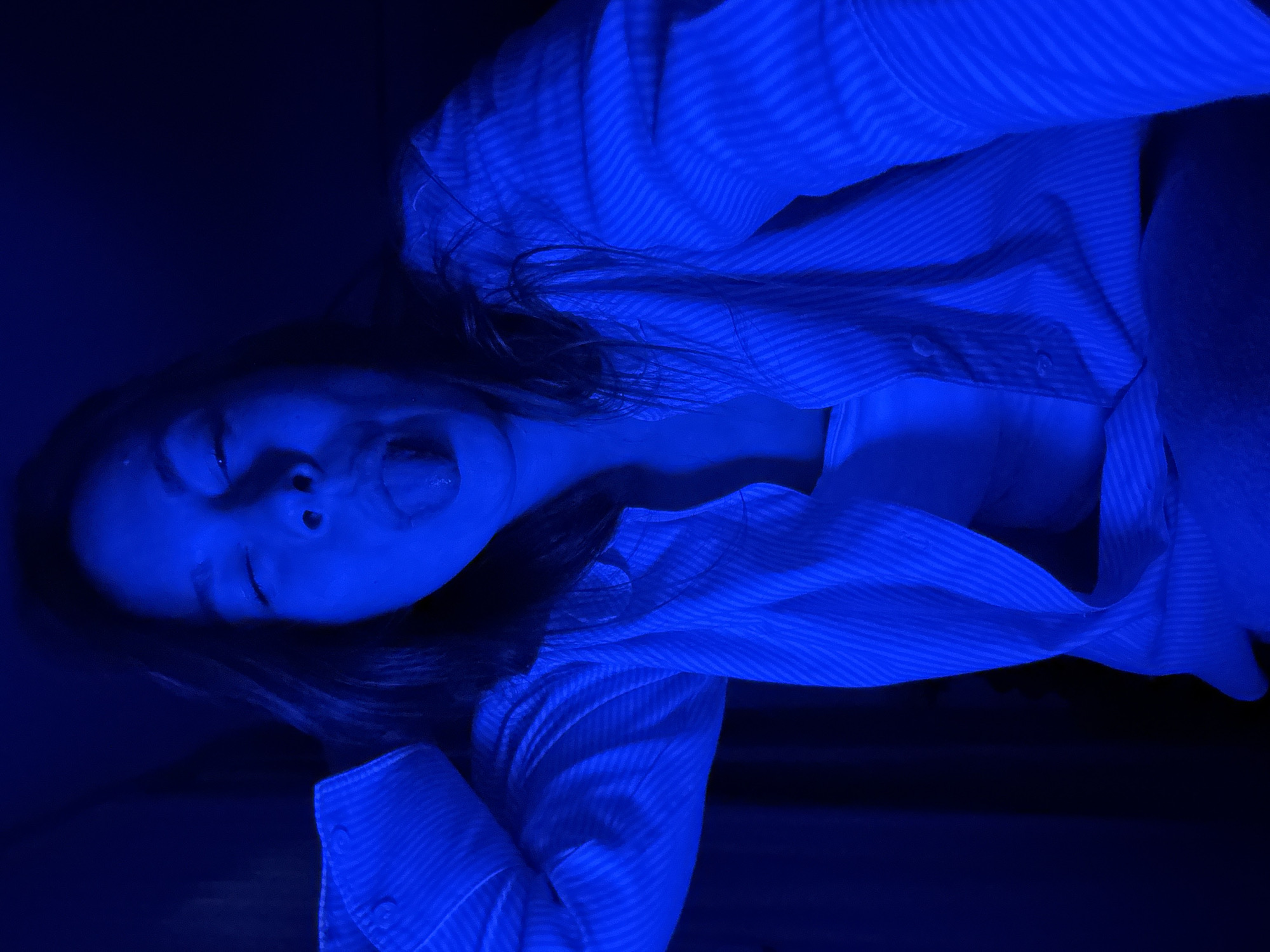 A person with long dark hair, wearing a striped shirt, is lying down, sticking their tongue out. The scene is lit with a blue light. There is no information to suggest this image was taken in the city of Budva, Montenegro.