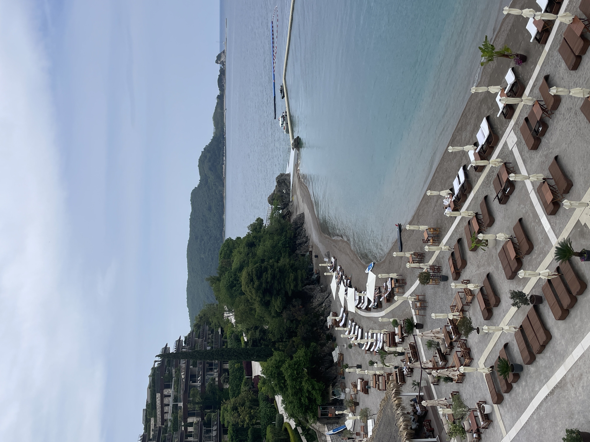 The image depicts a coastal scene, likely a beach resort, in Budva, Montenegro. The view is an elevated perspective, showing a coastline with light blue water and a beach area populated with numerous lounge chairs and umbrellas. The seating is arranged in organized rows on a gray surface that might be a concrete or paved area extending from the beach.
On the left side of the image, a modern multi-story building with extensive vegetation integrated into its design is visible, suggesting luxury accommodations. Lush green trees are intermixed with the building, contributing to a natural-urban blend.
The body of water stretches out toward the horizon, with a distant landmass barely visible in the upper left. A partially submerged strip of land extends into the water. There appear to be some boats and other structures at the edge of the water. The sky is overcast, creating soft lighting conditions.
People are visible occupying some of the lounge chairs near the shoreline. The overall scene is peaceful and suggests a high-end tourist destination.